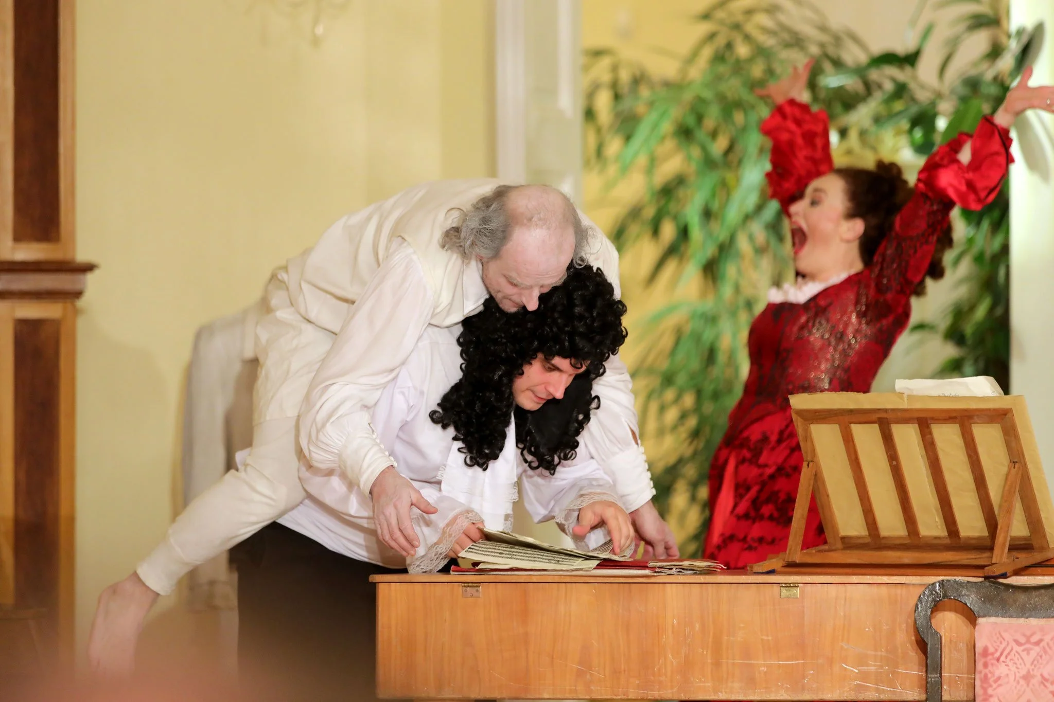 Drei Menschen in einem Theaterstück, zwei männlich in Kleidung aus der Vergangenheit, einer Frau in einem roten Kleid, dicht gedrängt um ein Klavier mit Notenblatt, während die Frau rechts vor Freude aufspringt.