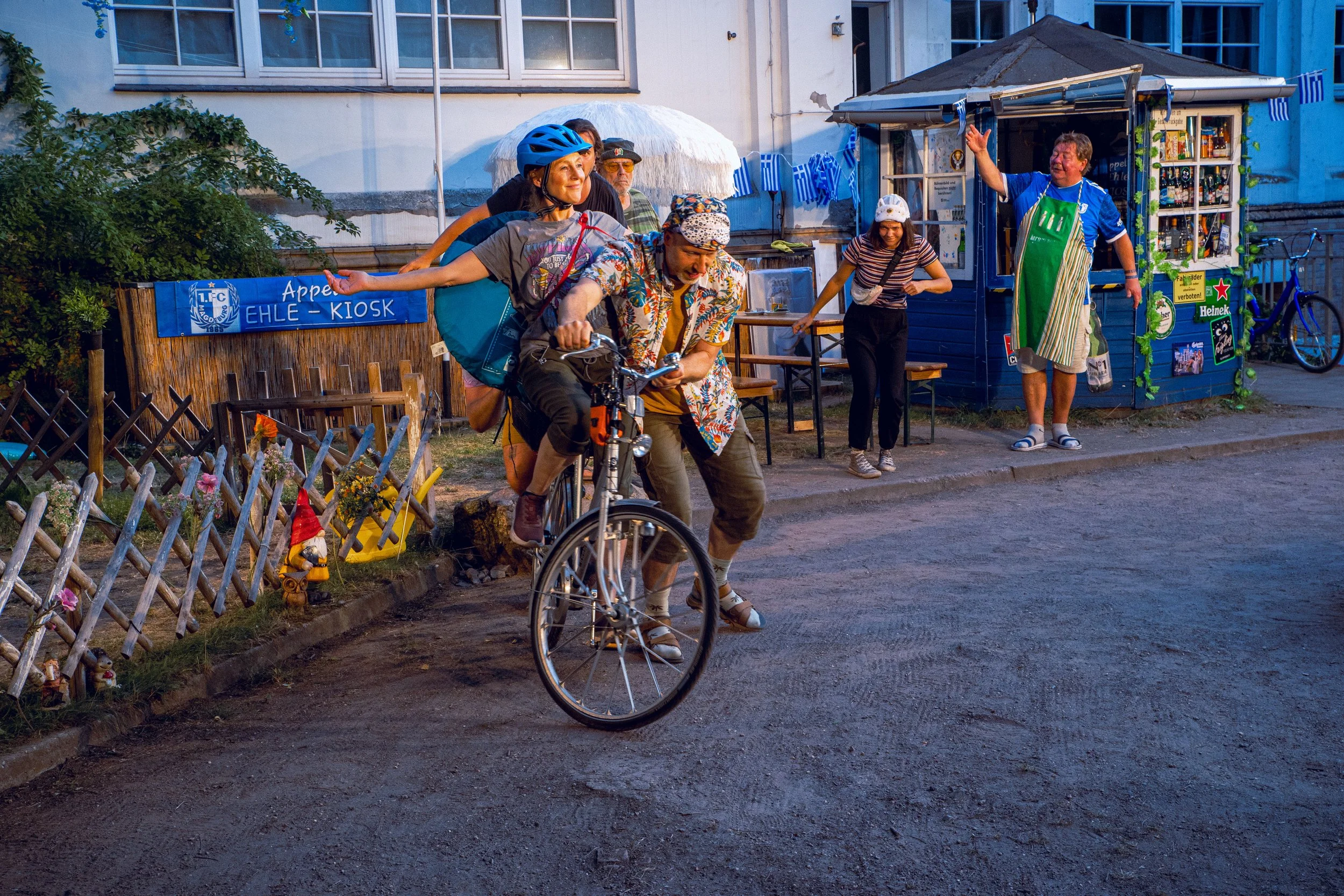 Menschen feiern auf einem Straßenfest mit einer kleinen Kaffeebude im Hintergrund, einige mit absurd großen Kopfbedeckungen. Es gibt eine Person, die auf einem Fahrrad sitzt und eine andere Person, die sie schiebt.