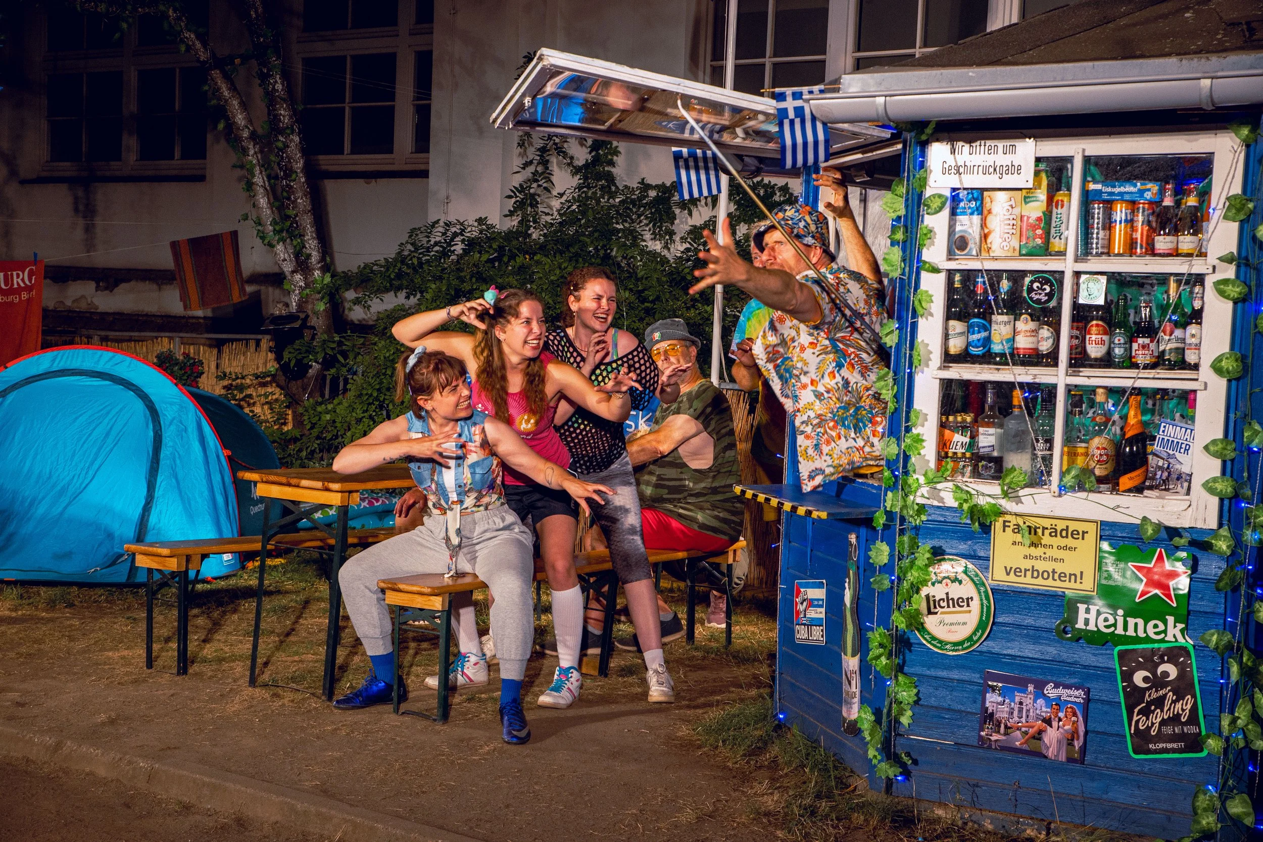 Gruppe von Menschen, die an einem Straßenhändlerstand lachen und Spaß haben, mit Zelten im Hintergrund, bei Nacht.