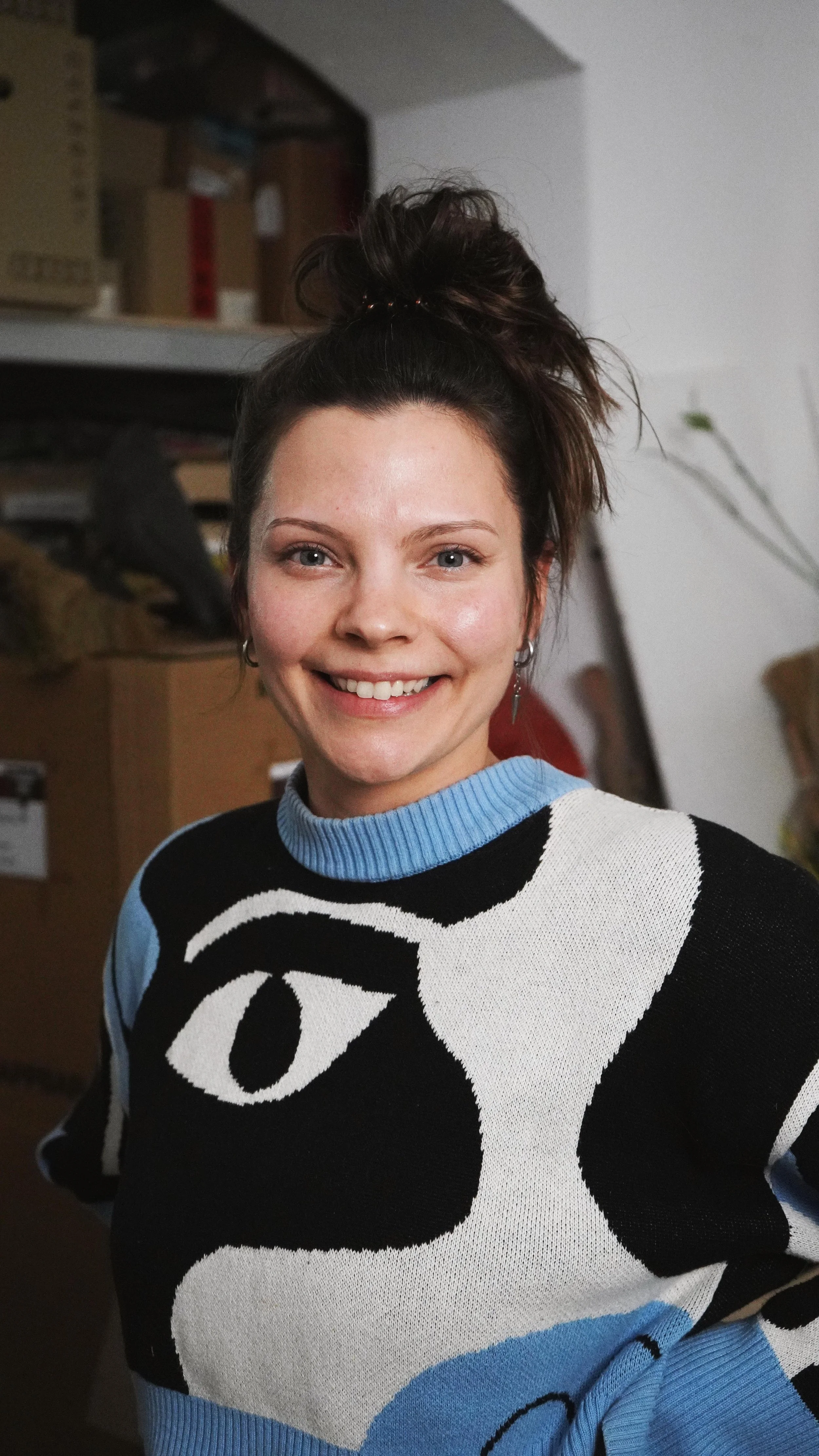 Junge Frau mit braunen Haaren in einem hohen Dutt, lächelnd, trägt ein schwarzes und weißes Musterpullover mit einem Katzenkopf-Design, steht in einem Raum mit Regal im Hintergrund.