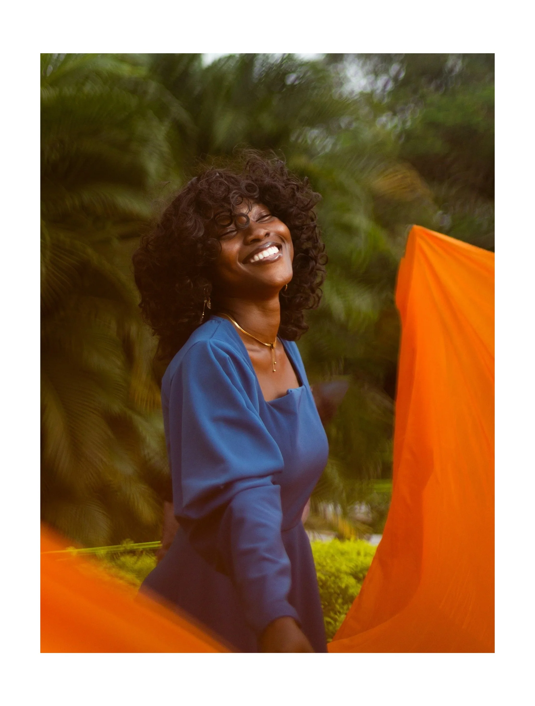 Happy, confident Black woman with blue blouse and gold jewelry in front of green leafy backdrop