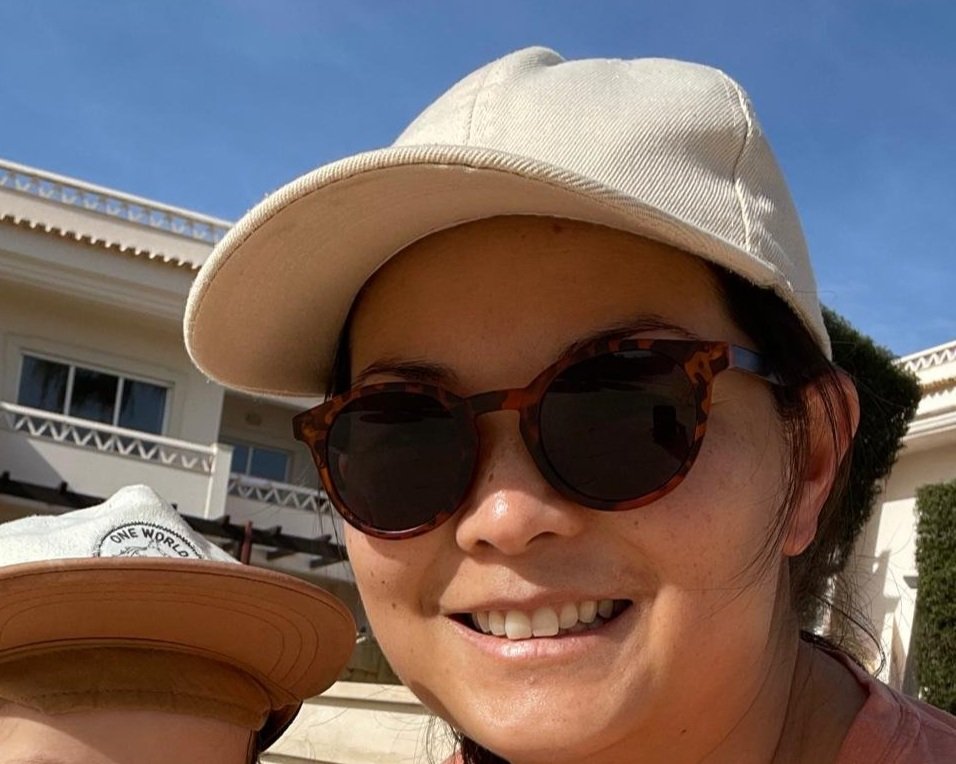 Close-up of a woman wearing tortoiseshell sunglasses, a beige cap, and smiling outdoors in front of a white building with balconies under a blue sky.