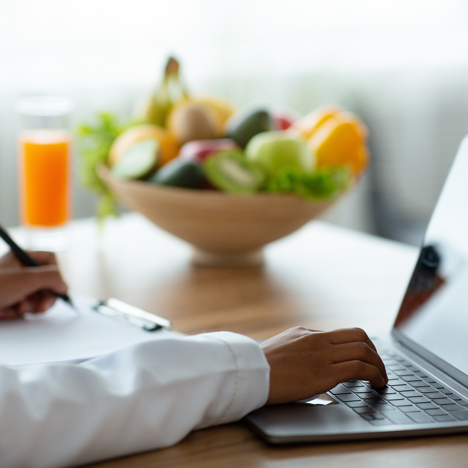 Person using a laptop at a wooden table, with a white bowl of mixed fruits and a glass of orange juice in the background.