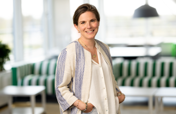 A smiling woman with short brown hair, wearing a light-colored blouse and a patterned jacket, standing in a modern, well-lit indoor space with green and white striped furniture in the background.