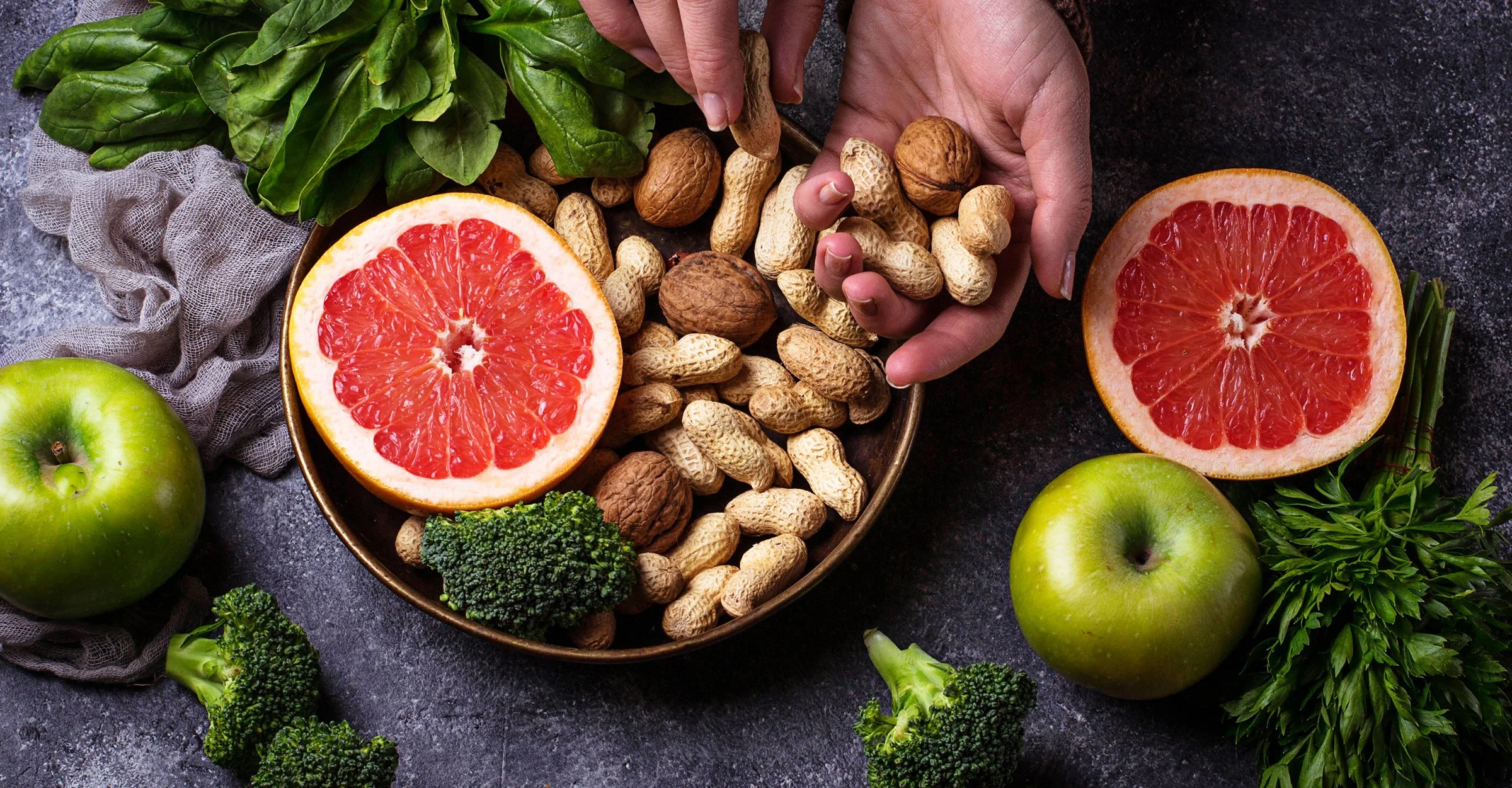A top-down view of a dark surface with a bowl of peanuts, two halves of grapefruit, green apples, broccoli, and fresh herbs.