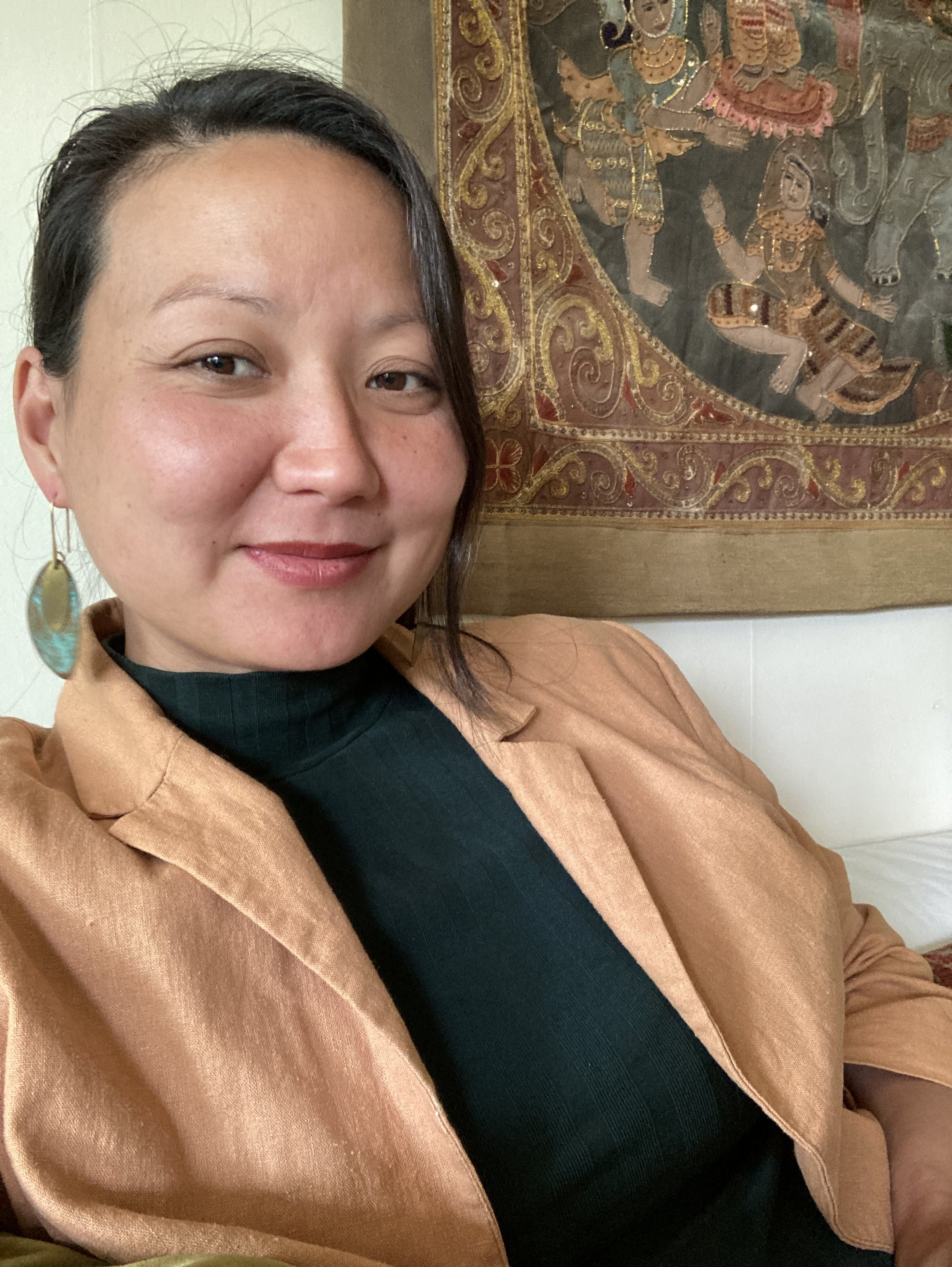A woman with short dark hair smiling, wearing a black turtleneck and a brown blazer, sitting in front of a traditional Asian tapestry with gold, red, and blue patterns and figures.