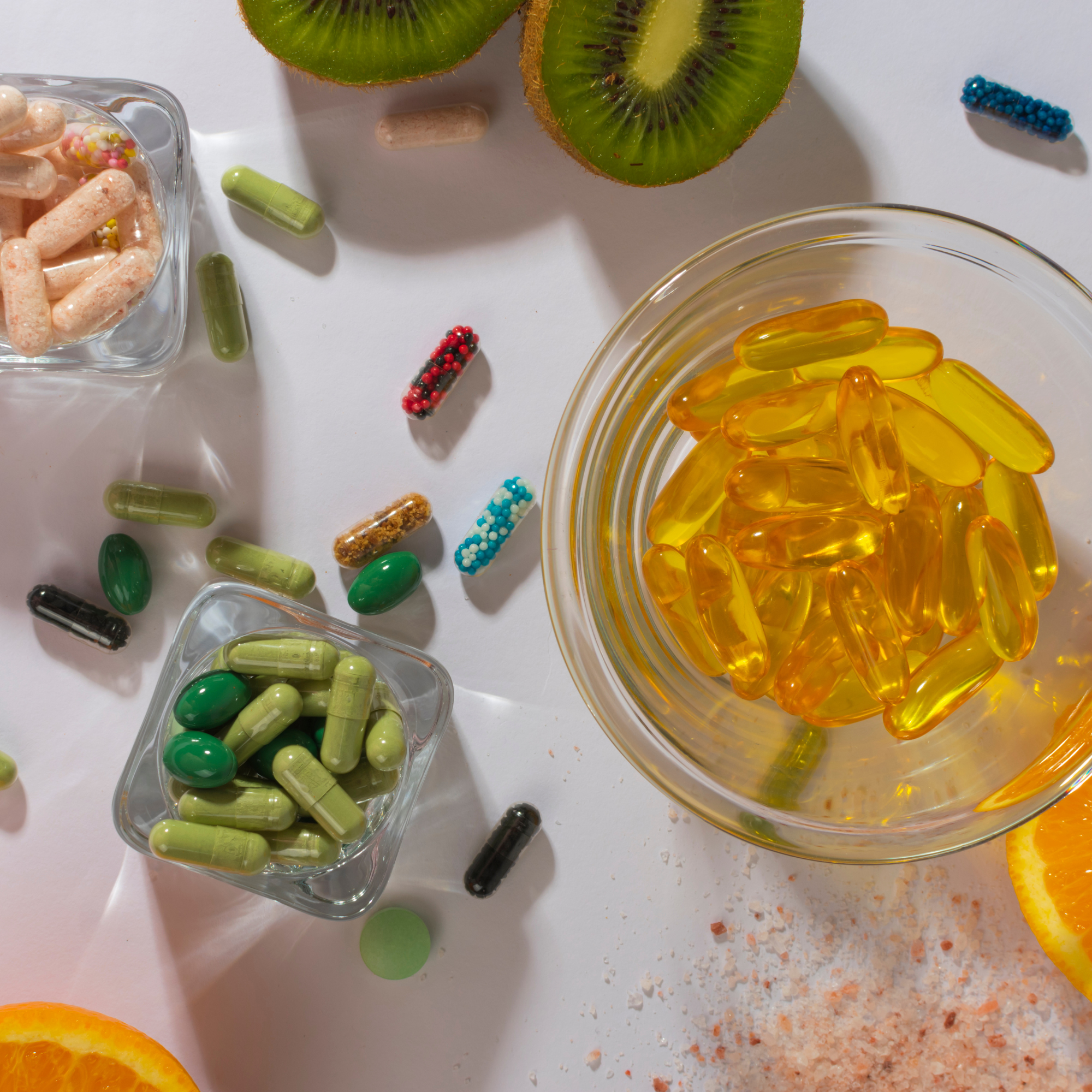 Assorted vitamins and supplements, including capsules and tablets, some in small bowls, with slices of kiwi, orange, and scattered salt on a white surface.
