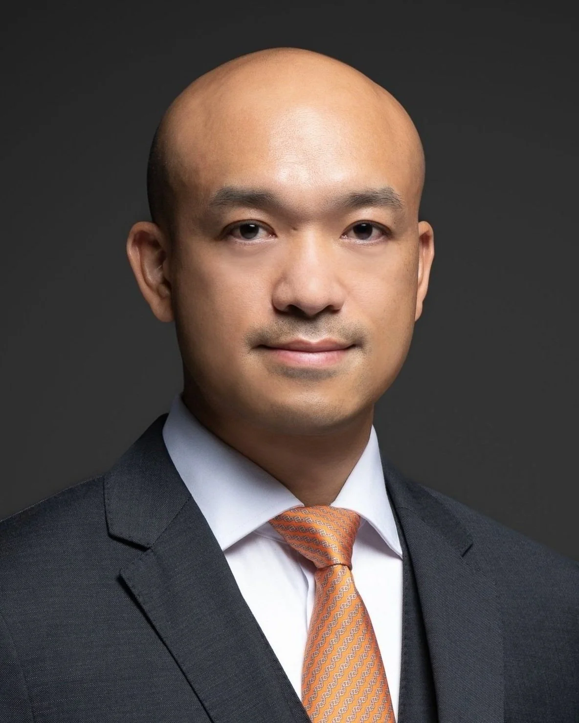 Professional headshot of a man with a shaved head, wearing a dark suit, white shirt, and orange tie, against a dark background.