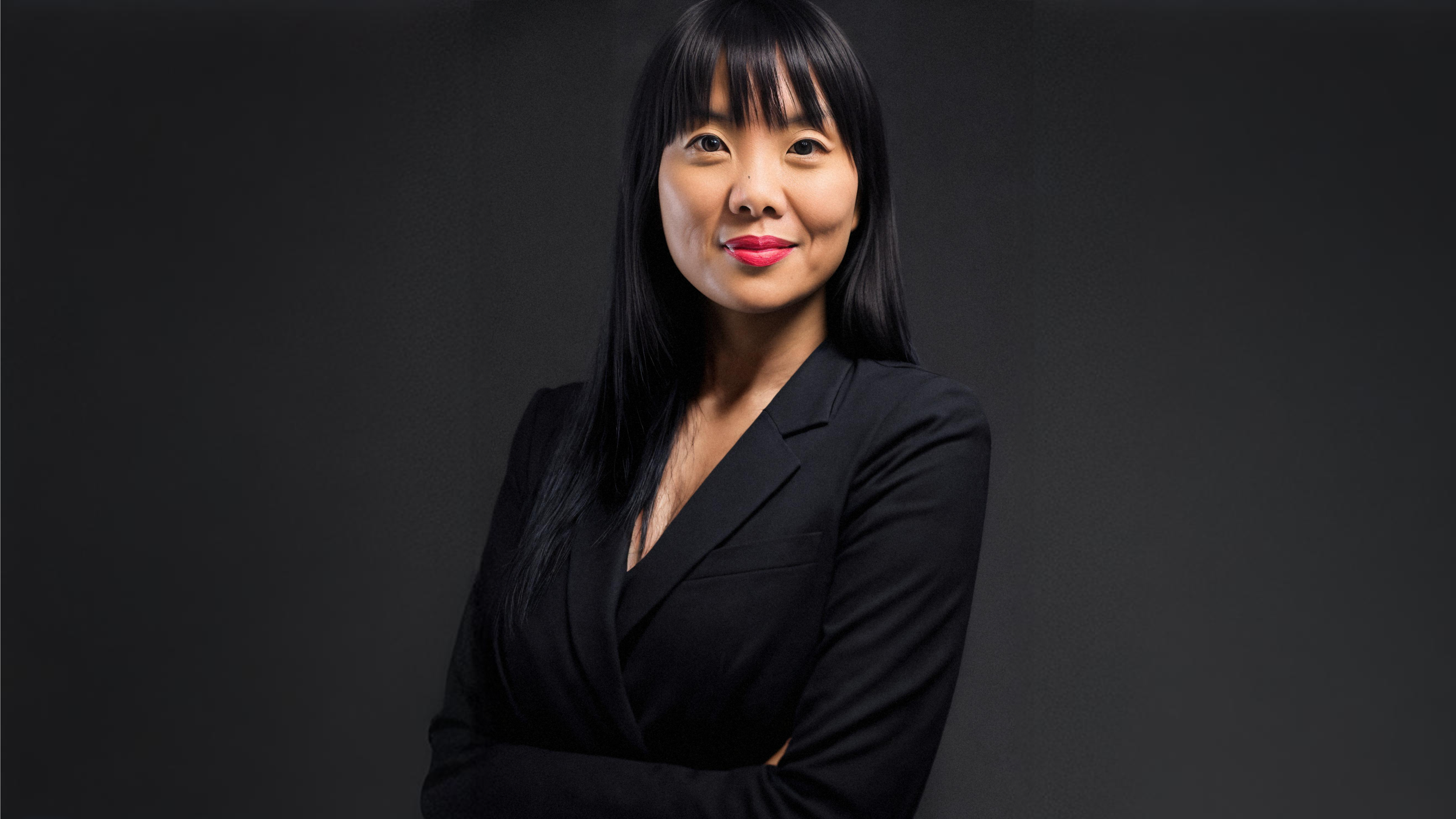 A portrait of an Asian woman with long black hair, wearing a black blazer, smiling against a dark gray background.