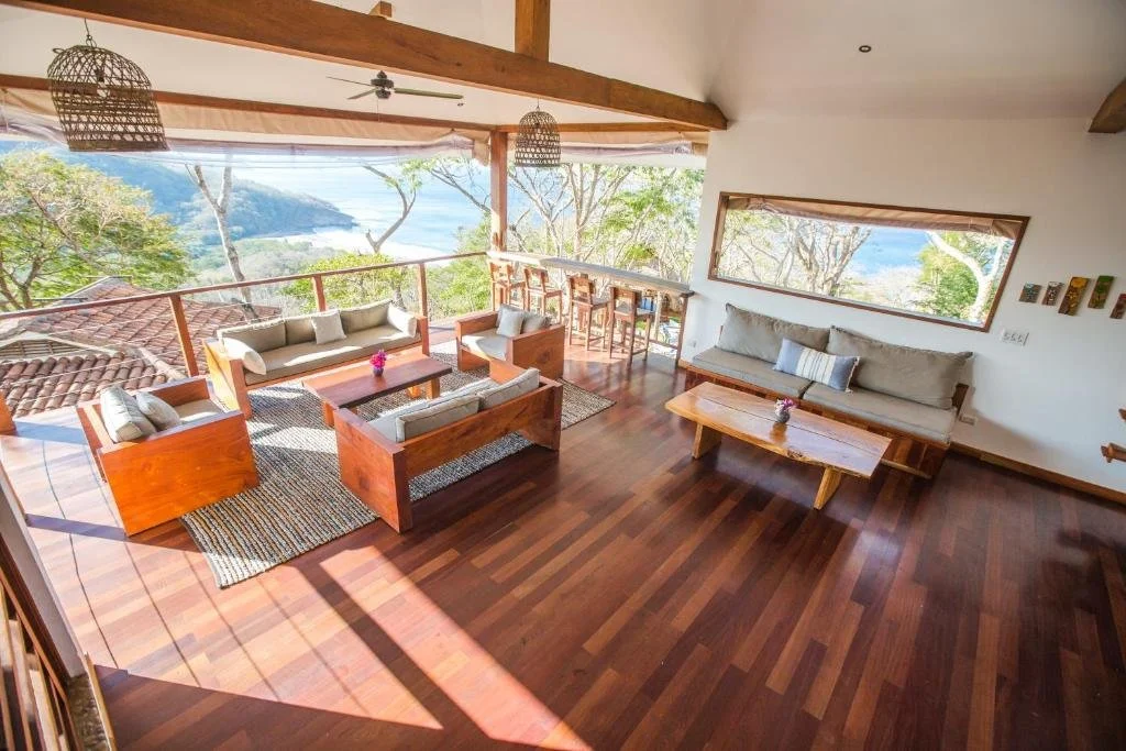 Living room with wooden furniture, large windows showing trees and a body of water, and a polished wooden floor.