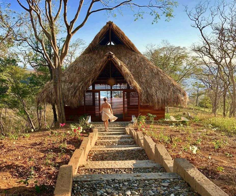 A woman in a white dress walking up a stone and wood staircase towards a thatched-roof hut in a natural, wooded setting with trees and plants.