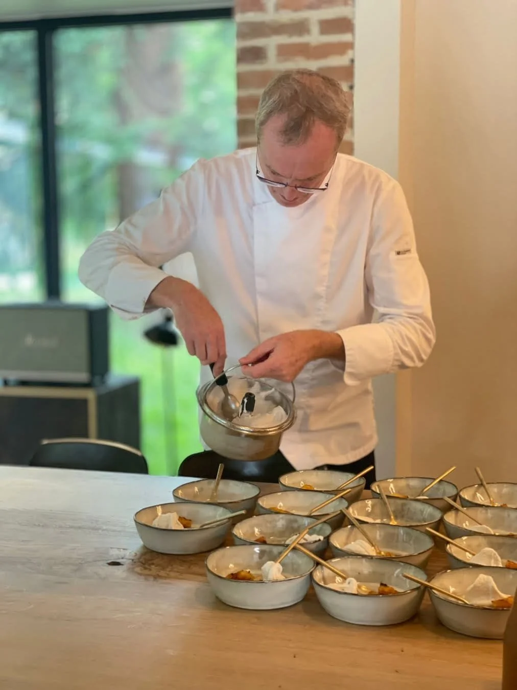 A chef wearing glasses and a white coat is preparing desserts in a bowl, with several finished bowls with gold spoons on a wooden table.