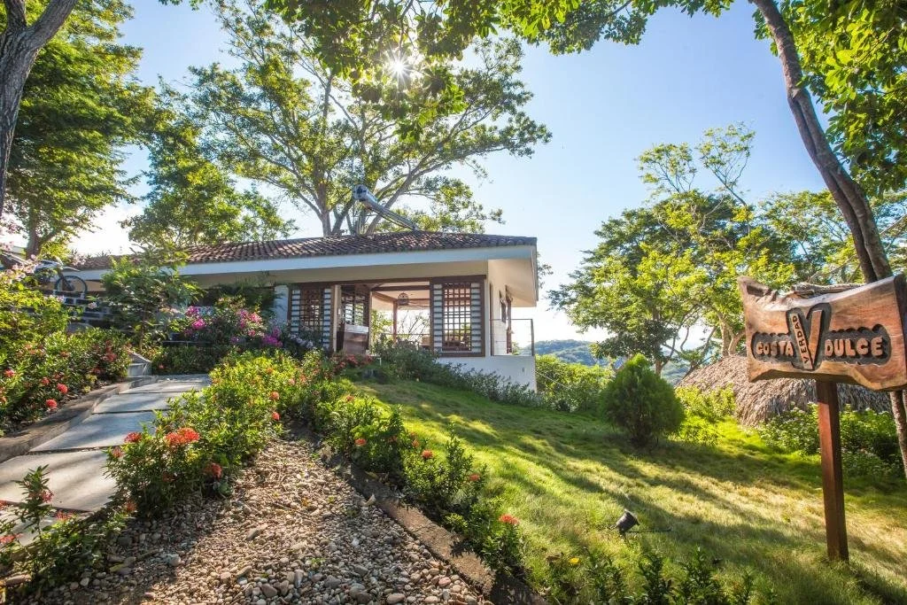 A hillside garden with colorful flowers, a paved pathway leading to a small house with a tiled roof, surrounded by lush green trees, with a wooden sign that reads "Costa Verda Dulce" and sunny sky in the background.