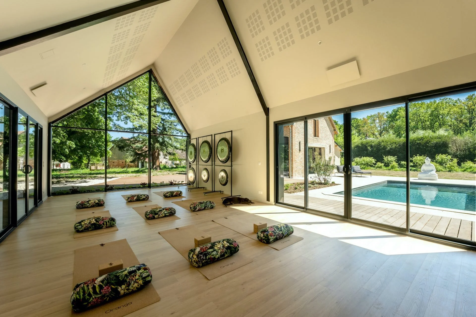 Indoor yoga or meditation studio with wooden floor, floral pillows on mats, and large windows overlooking a backyard with a swimming pool, trees, and a Buddha statue.