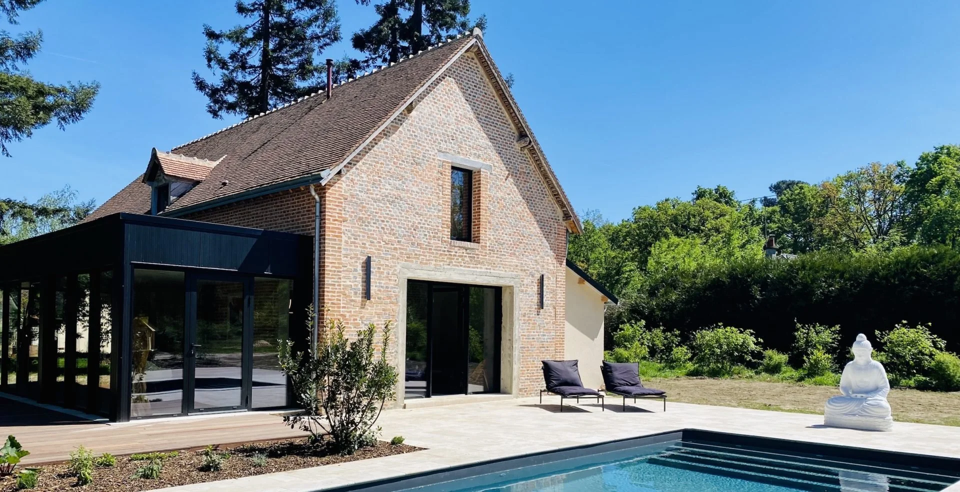 A brick house with a modern glass extension, outdoor patio with two lounge chairs, a statue of Buddha near a swimming pool, surrounded by greenery and trees under a clear blue sky.