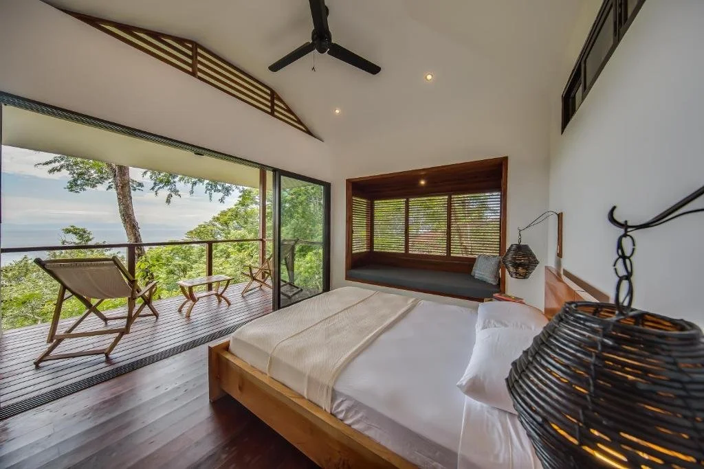 A bedroom with a large glass door opening to a balcony overlooking trees and water, featuring a wooden bed, chairs, and a built-in seating area with wooden blinds.