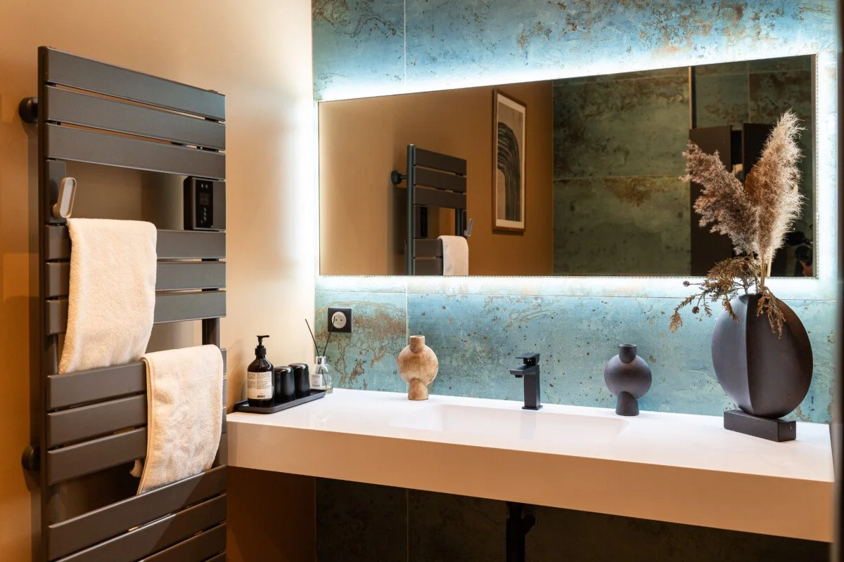 Modern bathroom with a large mirror, black faucet, decorative vases, dried plants in a black vase, and a gray heated towel rack with towels.