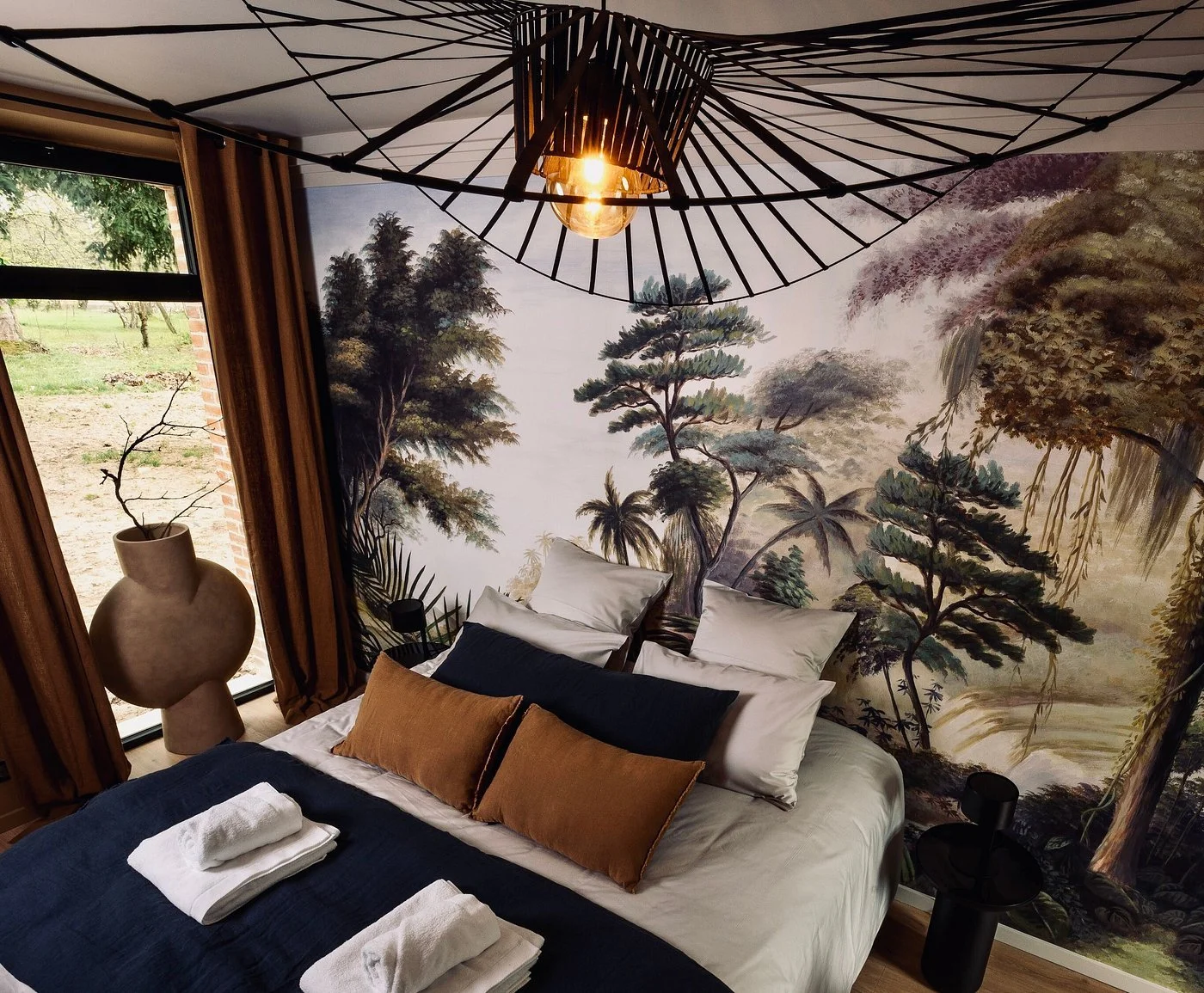 A bedroom with a wall mural of a jungle landscape scene, a window with curtains, a large decorative vase with a branch, and neatly made bed with white, navy blue, and brown pillows and towels.