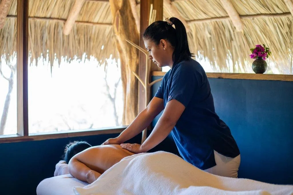 Massage therapist giving a back massage to a client in a tropical-style room with thatched roof and flowers on a shelf.