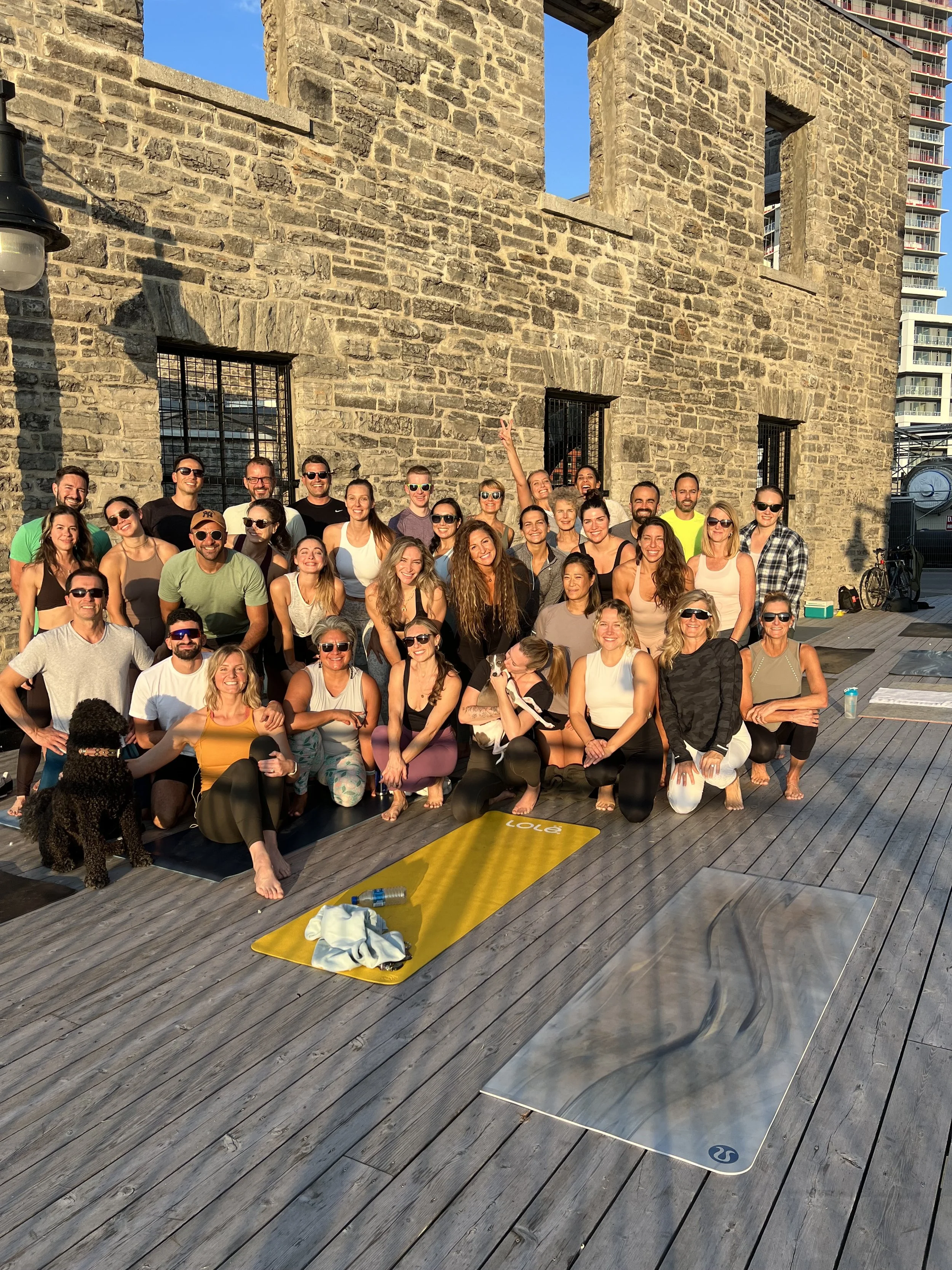 A large group of people posing outdoors on a wooden deck in front of a stone building, with some yoga mats and a black dog sitting in front. The group is smiling and enjoying sunny weather.