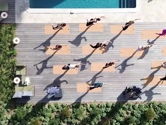 Top-down view of people practicing yoga on yoga mats outdoors, with shadows cast on a wooden deck near a pool, surrounded by greenery.