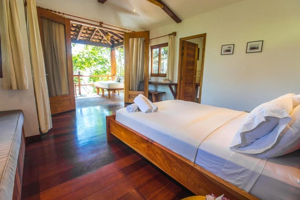 A cozy bedroom with a wooden bed, recent linens, and a view of a screened balcony with outdoor seating amid lush greenery.