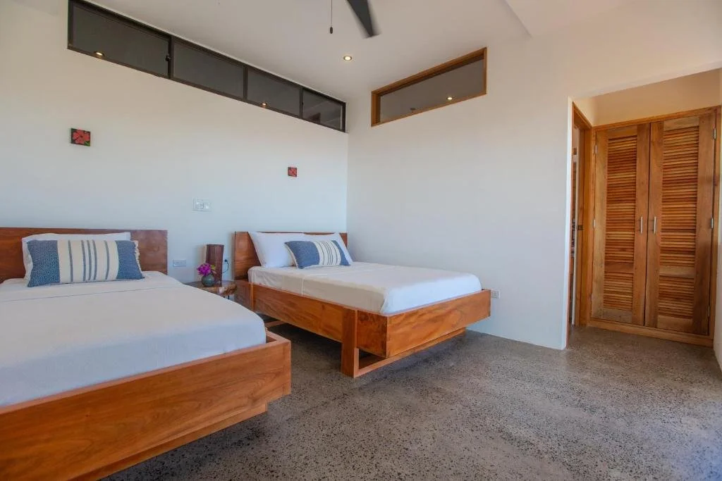 A bedroom with two wooden framed beds, white bedding, and striped pillows. A bedside table with flowers and a lamp, wooden closet doors, and small decorative wall art. Carpeted floor and a ceiling fan.