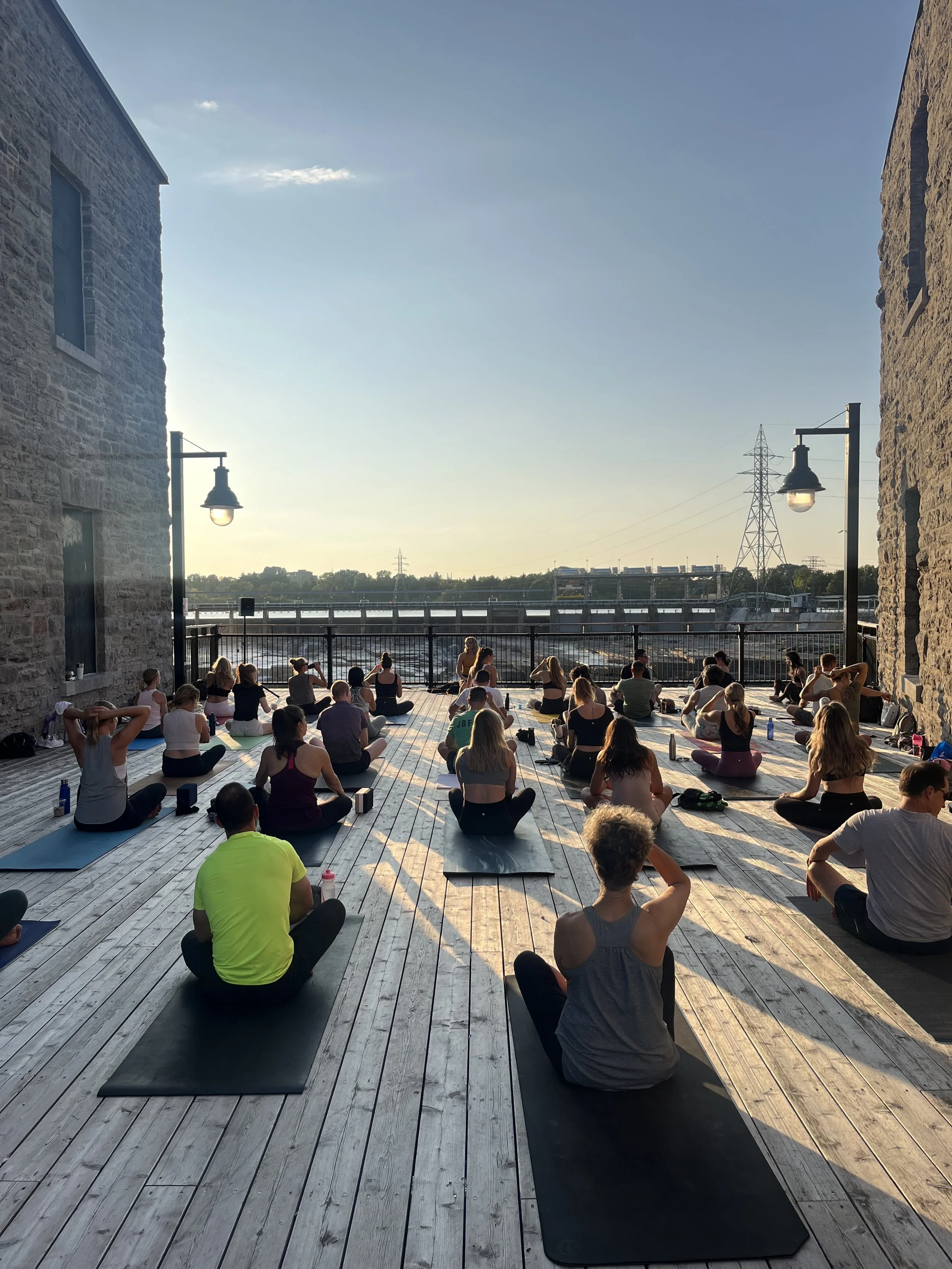 Group of people participating in outdoor yoga class during sunset on a wooden deck between stone buildings.