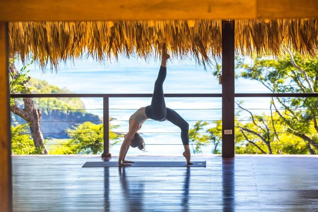 Person practicing a yoga pose on a wooden deck with ocean and trees in the background.