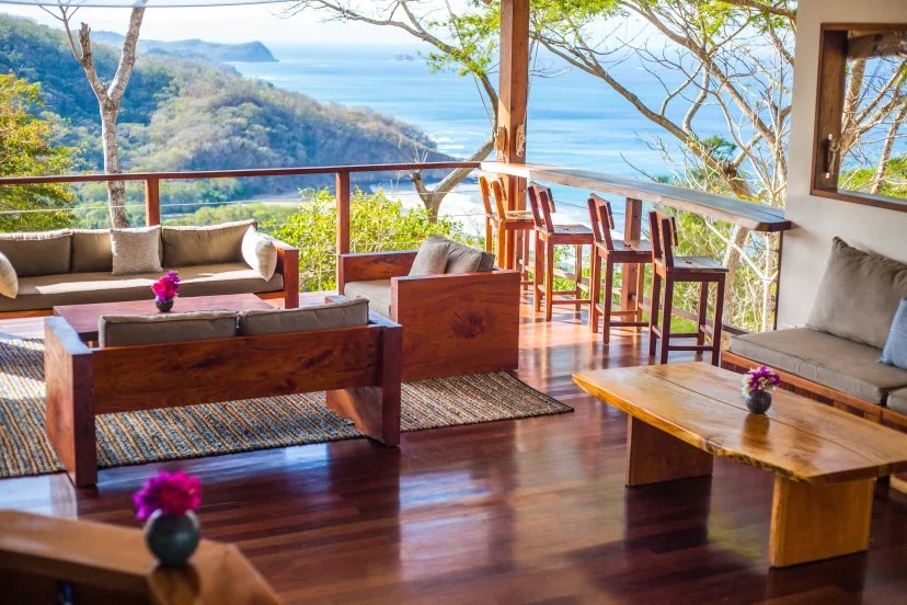 Living room with wooden furniture, beige sofas with pillows, and a view of trees, ocean, and sky outside through large windows and open balcony