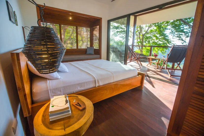 A cozy bedroom with a large bed, a hanging wicker lamp, a wooden nightstand with books, and a balcony with outdoor chairs overlooking trees and water.