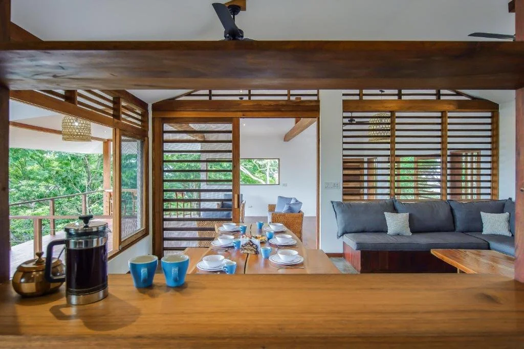 Interior of a modern home featuring a dining area with a wooden table set for four, a gray sofa with white pillows, and wooden slat partitions, with a lush green outdoor view through large windows.