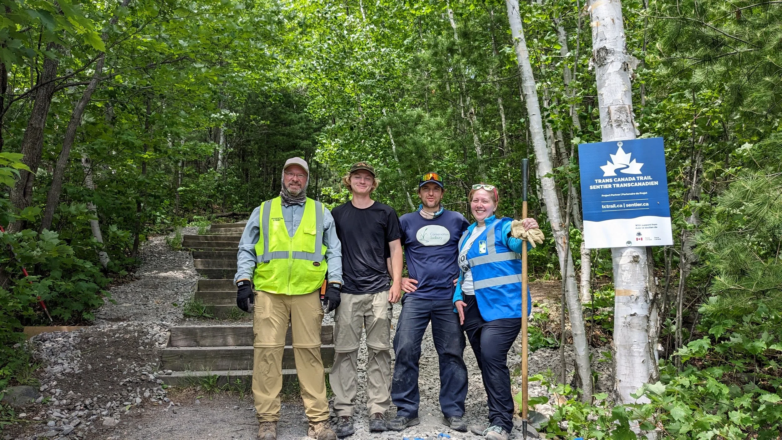 Four people standing on a trail in a forest, smiling, with a blue sign on a tree that reads "Trans Canada Trail." The group includes two men wearing high-visibility vests and two women in outdoor gear, all appearing to be participating in a trail maintenance or conservation project.