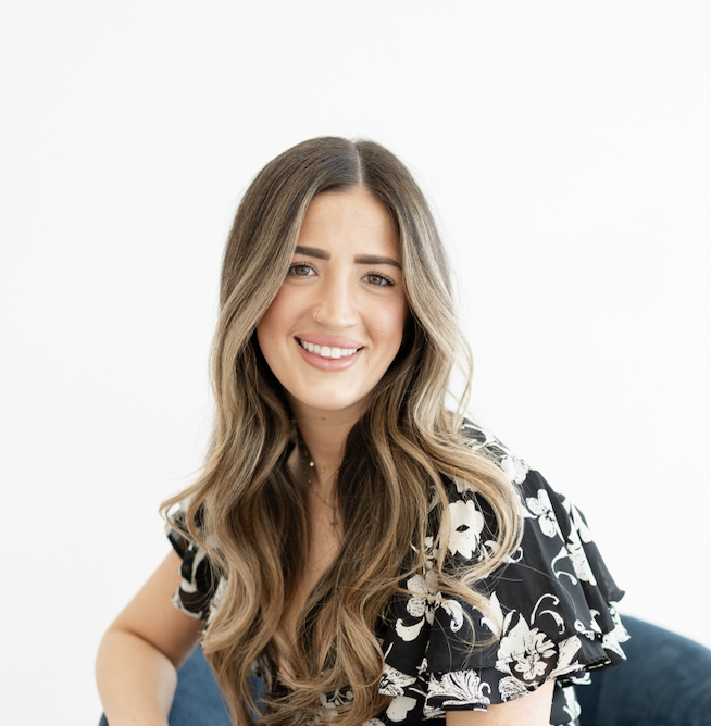 A young woman with long, wavy brown hair, smiling while sitting on a dark blue chair in front of a plain white background.