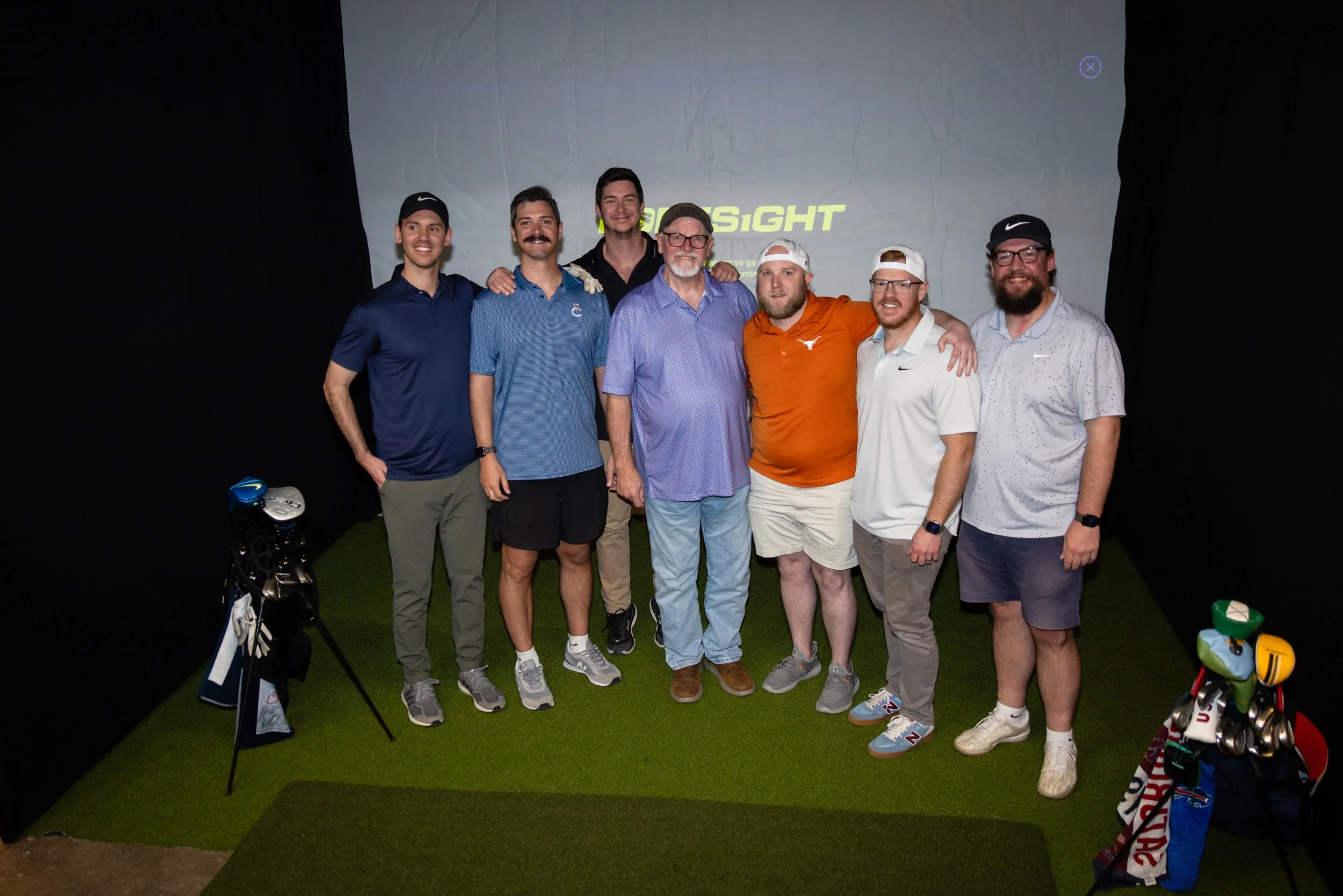 Group of seven men posing in an indoor golf simulator room, some with golf clubs and golf bags, with a screen projecting a golf course view behind them.