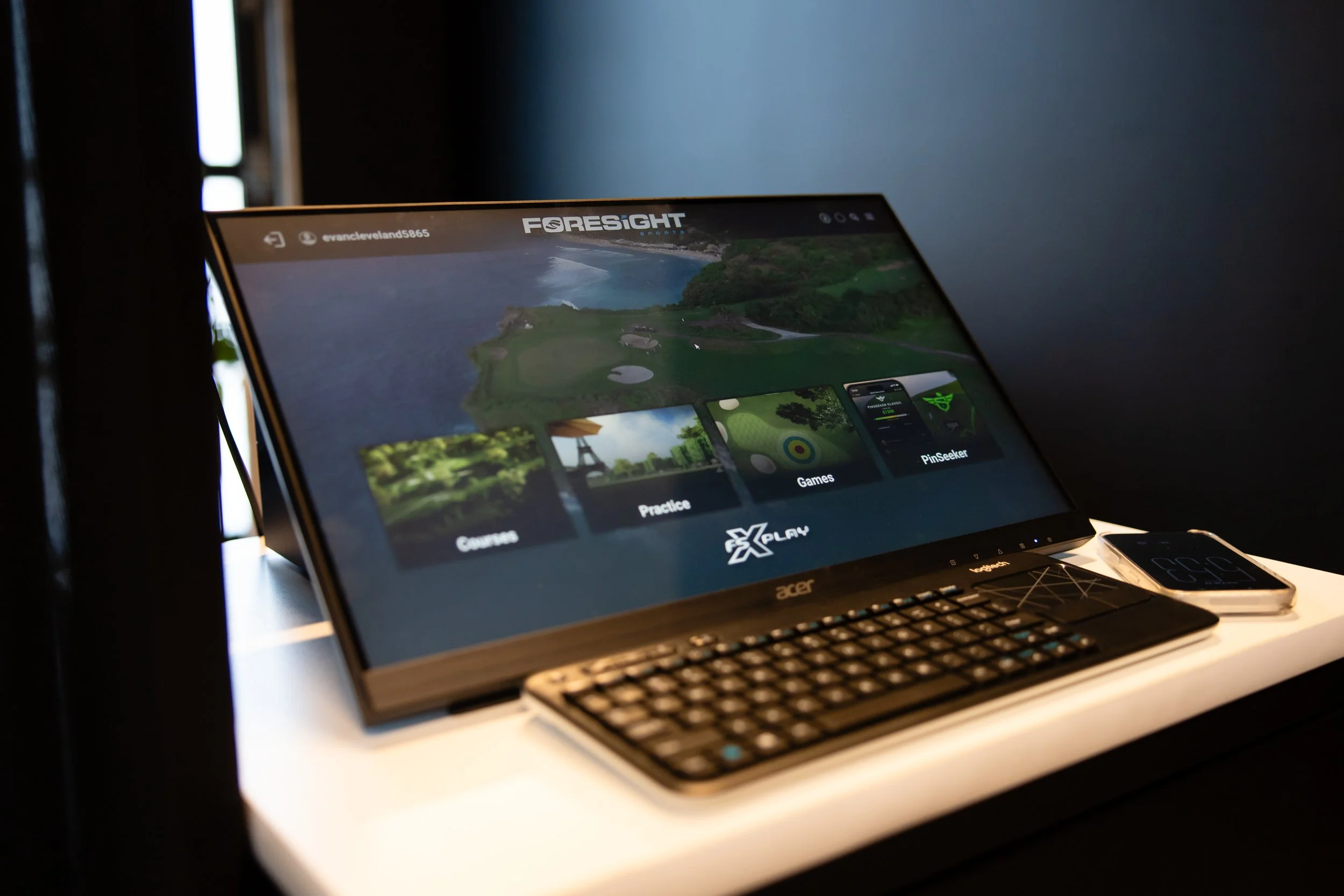 A computer monitor displaying the Foresight Sports app interface, with options for courses, practice, games, and PinSeeker, placed on a white desk alongside a wireless keyboard and a smartphone showing a golf-related app.