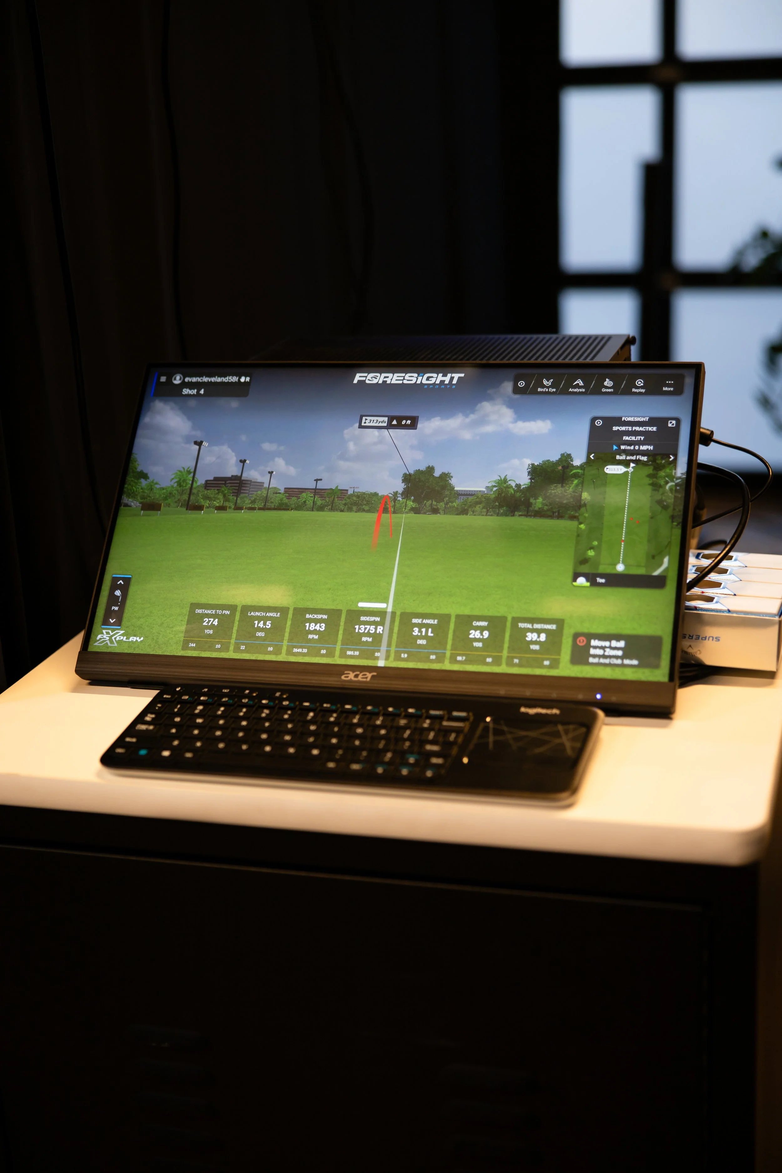 Golf simulation software on a monitor displaying a golf shot trajectory on a field, with a keyboard placed in front, on a white desk, near a window.