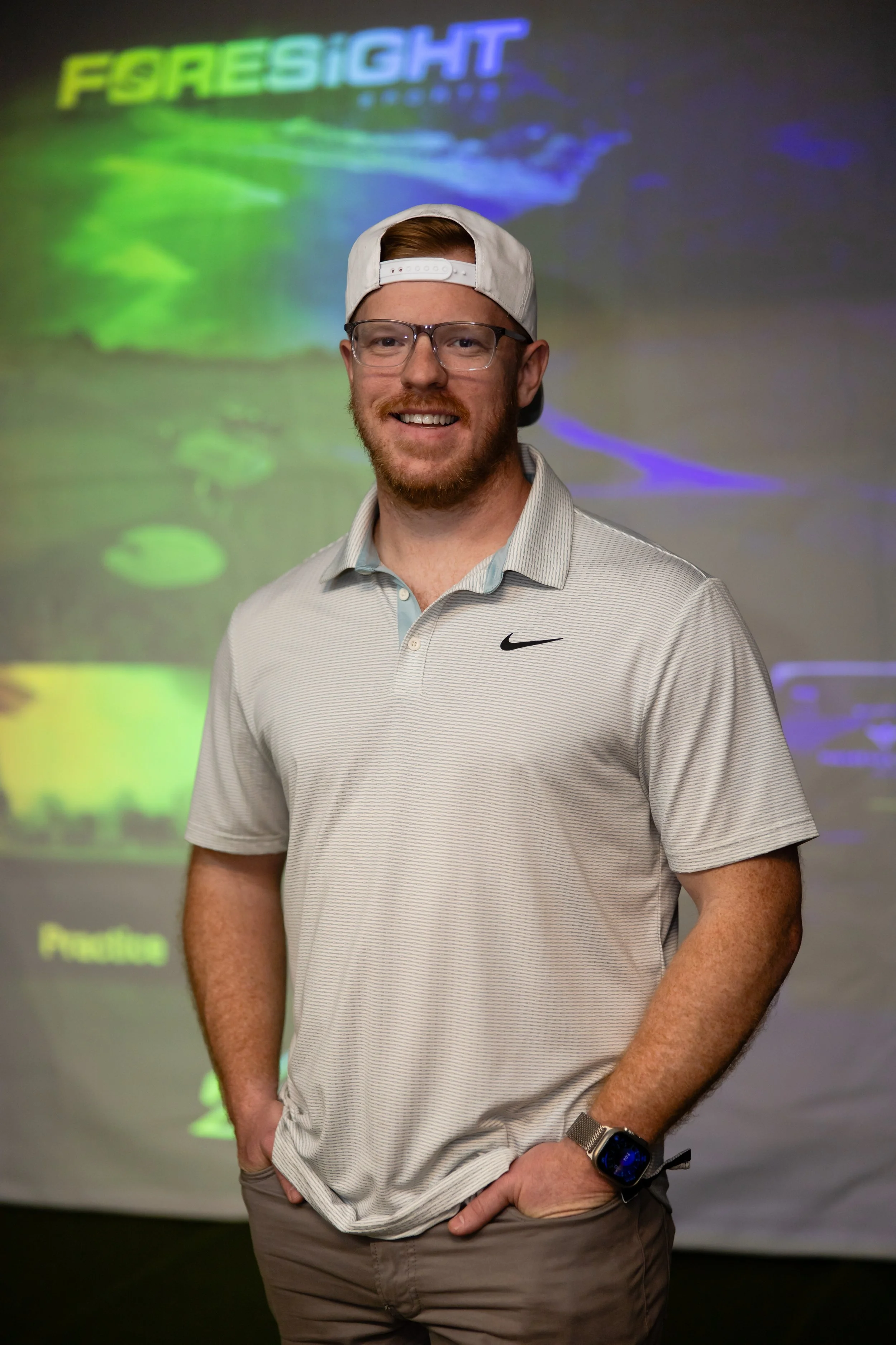 A smiling man with glasses, a white cap worn backwards, a gray polo shirt, standing in front of a golf simulator.