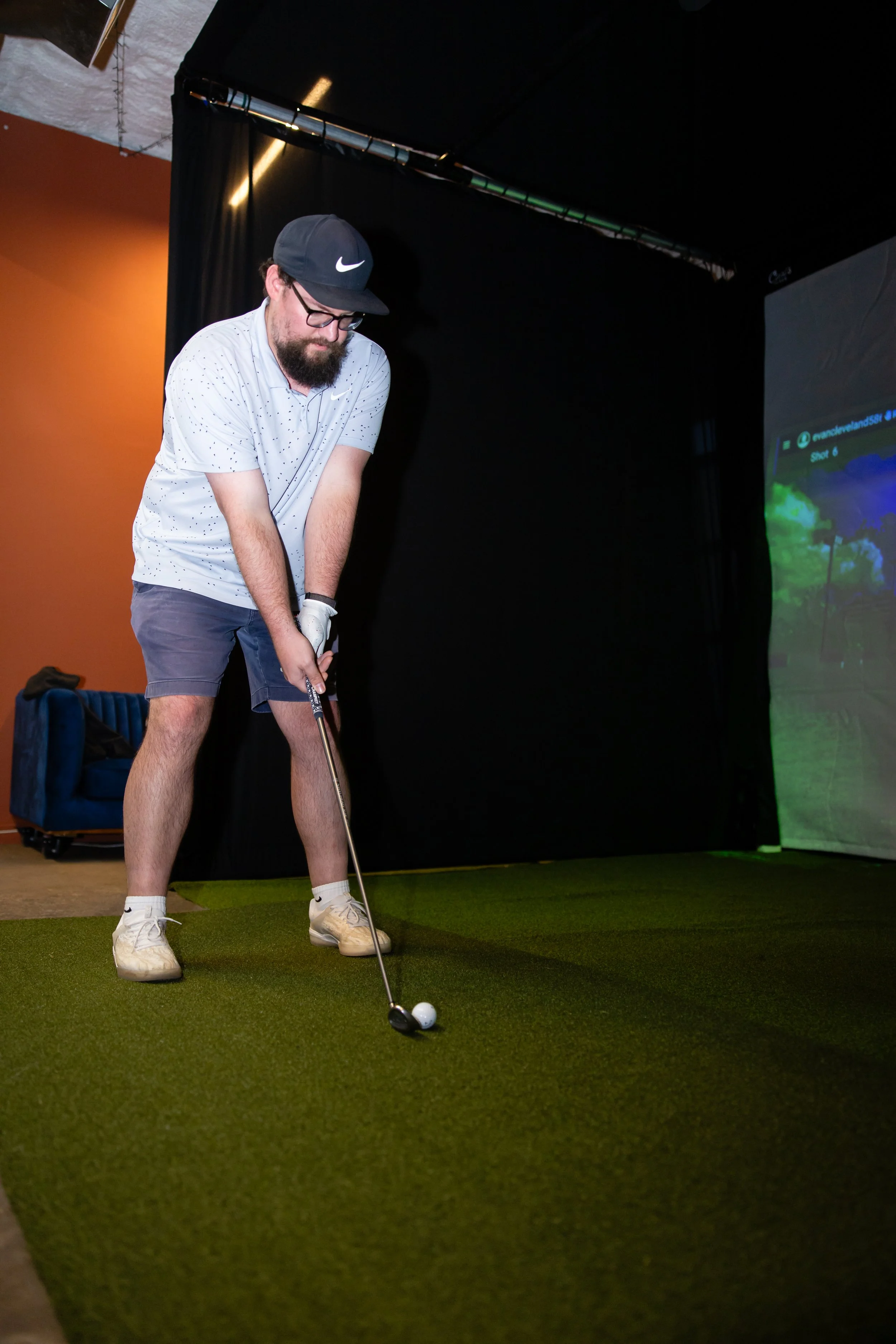 A man wearing a dark blue Nike cap, glasses, a white dotted polo shirt, gray shorts, and white golf shoes, is preparing to hit a golf ball inside a room with artificial turf and black curtains.