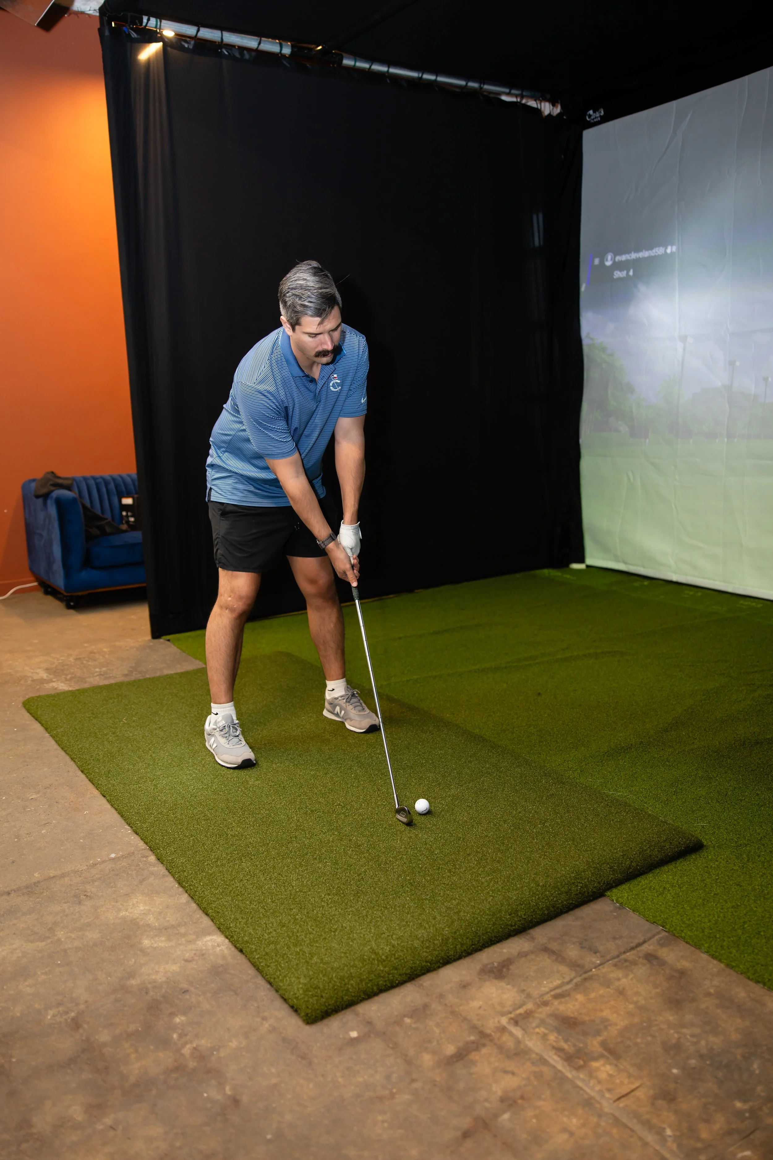 Man practicing indoor golf in a simulation room with green turf and digital screen