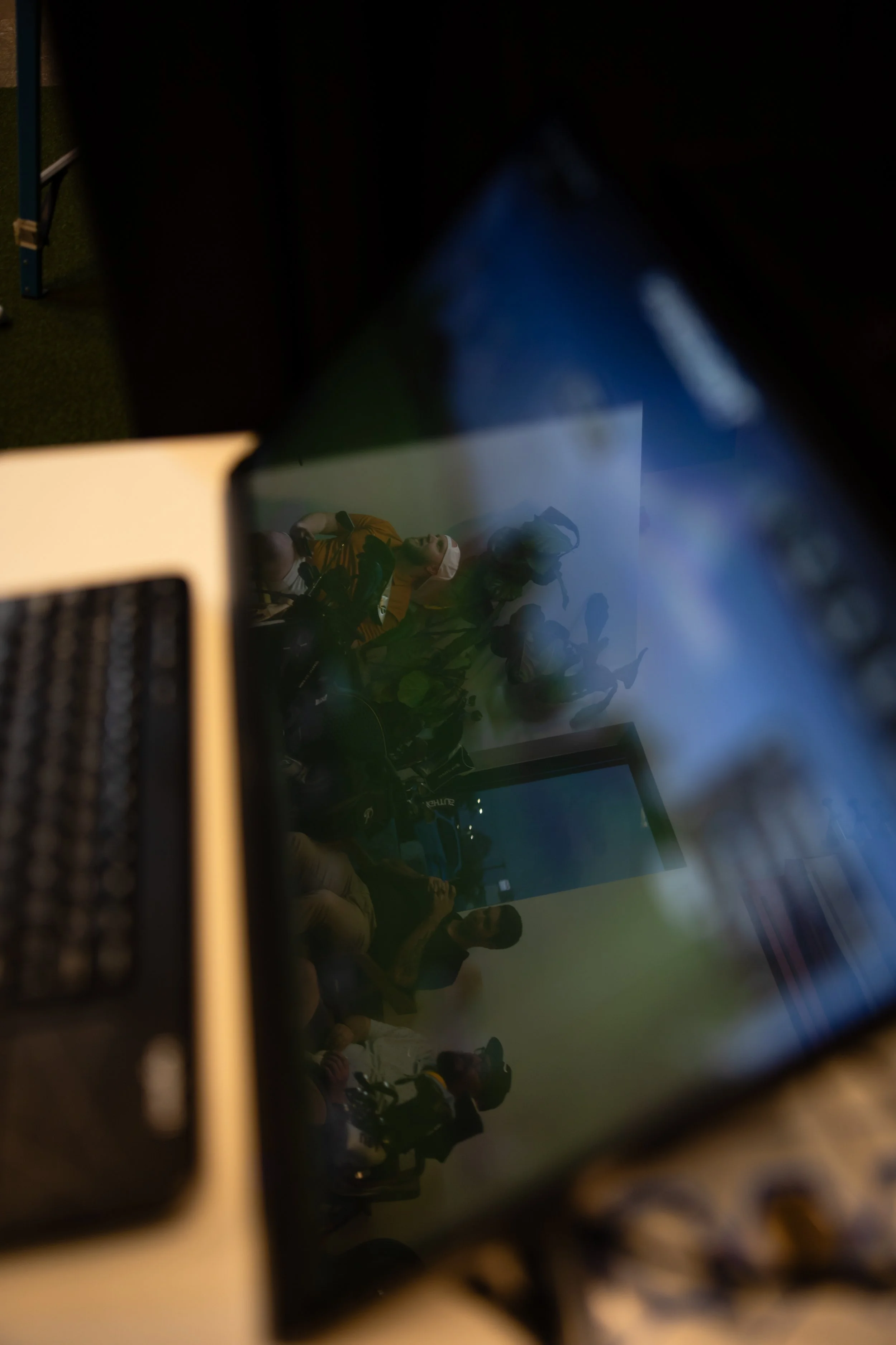 A group of people taking a selfie, viewed through a reflective surface, with some wearing helmets and vests, and a screen or monitor visible in the background.