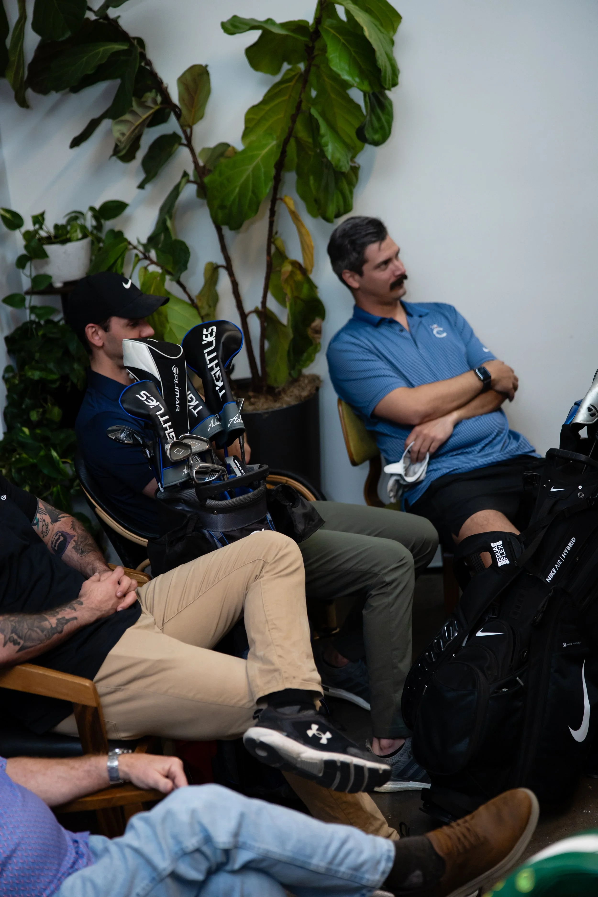 A group of men sitting in a room, some with golf clubs, with large potted plants in the background.