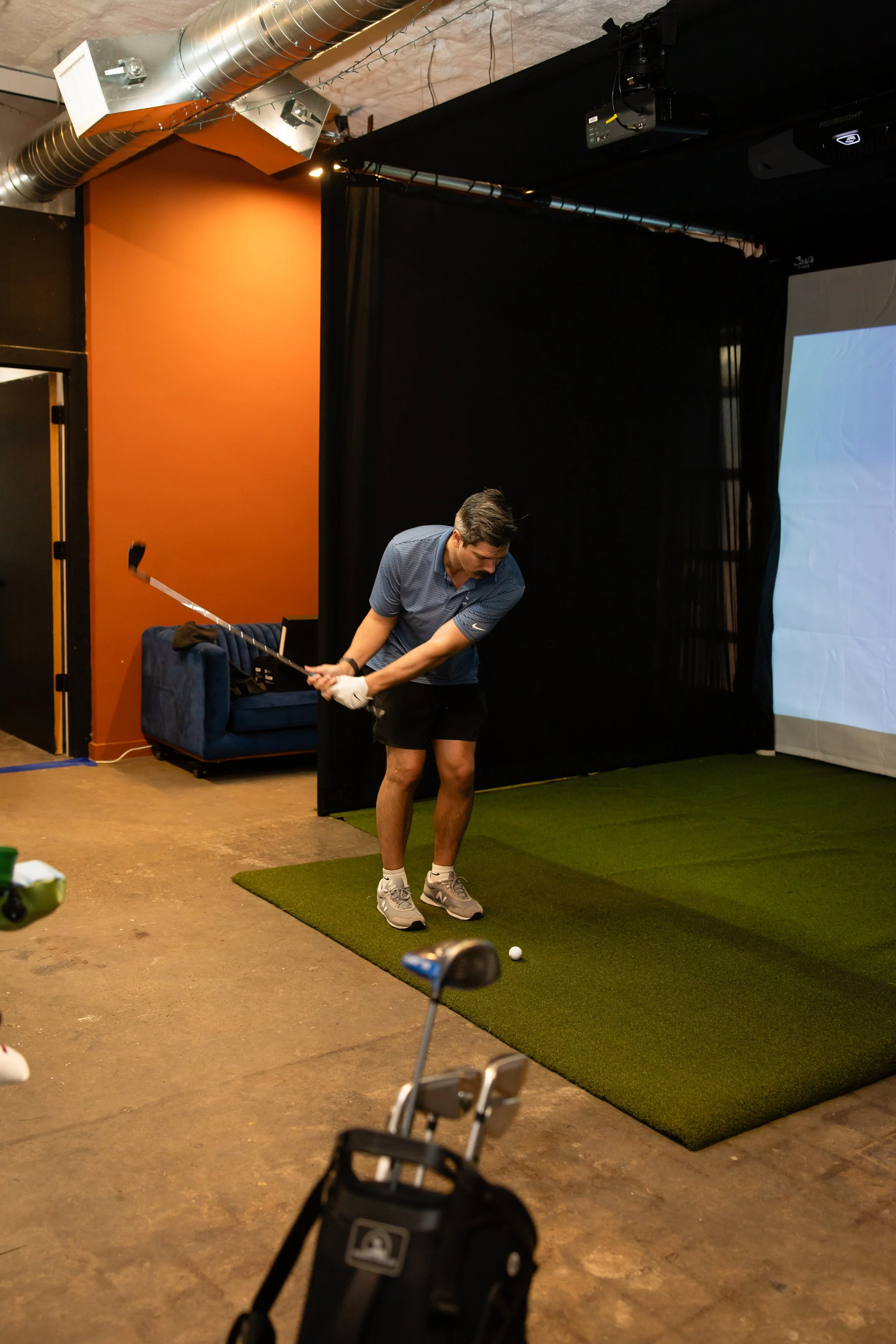 Young man practicing golf indoors, preparing to hit a ball on a small artificial turf mat, with golf clubs in a golf bag nearby.
