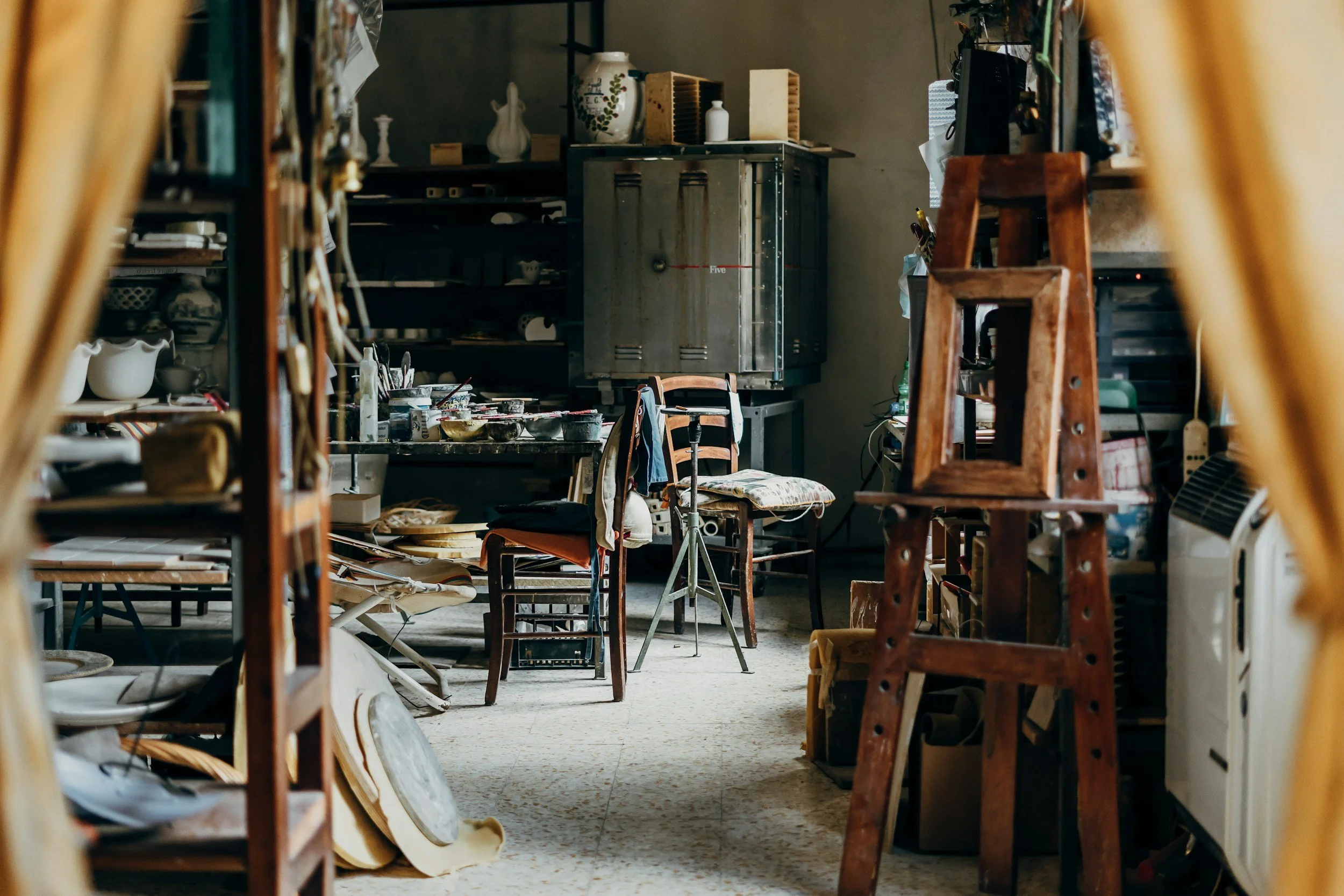 Artist’s studio filled with easels, chairs, shelves, and art supplies, viewed through a partially open doorway.