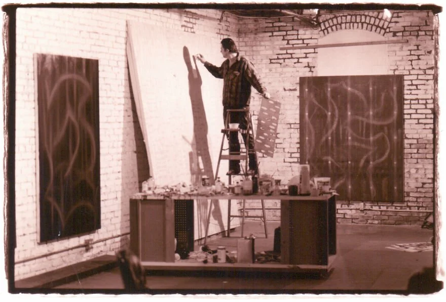 An artist is standing on a stepladder, working on a large white canvas in an art studio with brick walls. There are two other abstract paintings hanging on the walls and various art supplies on a table below.