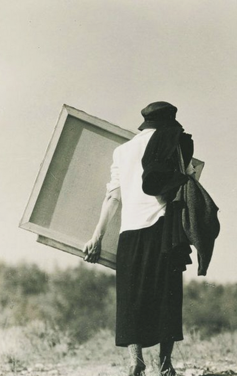 Black-and-white photograph of an artist outdoors carrying a large blank canvas, with a dark hat and coat draped over one shoulder.