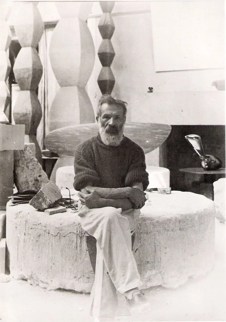 Black and white photo of a man with a beard sitting on a large stone, surrounded by art or sculpture pieces in a studio or gallery.