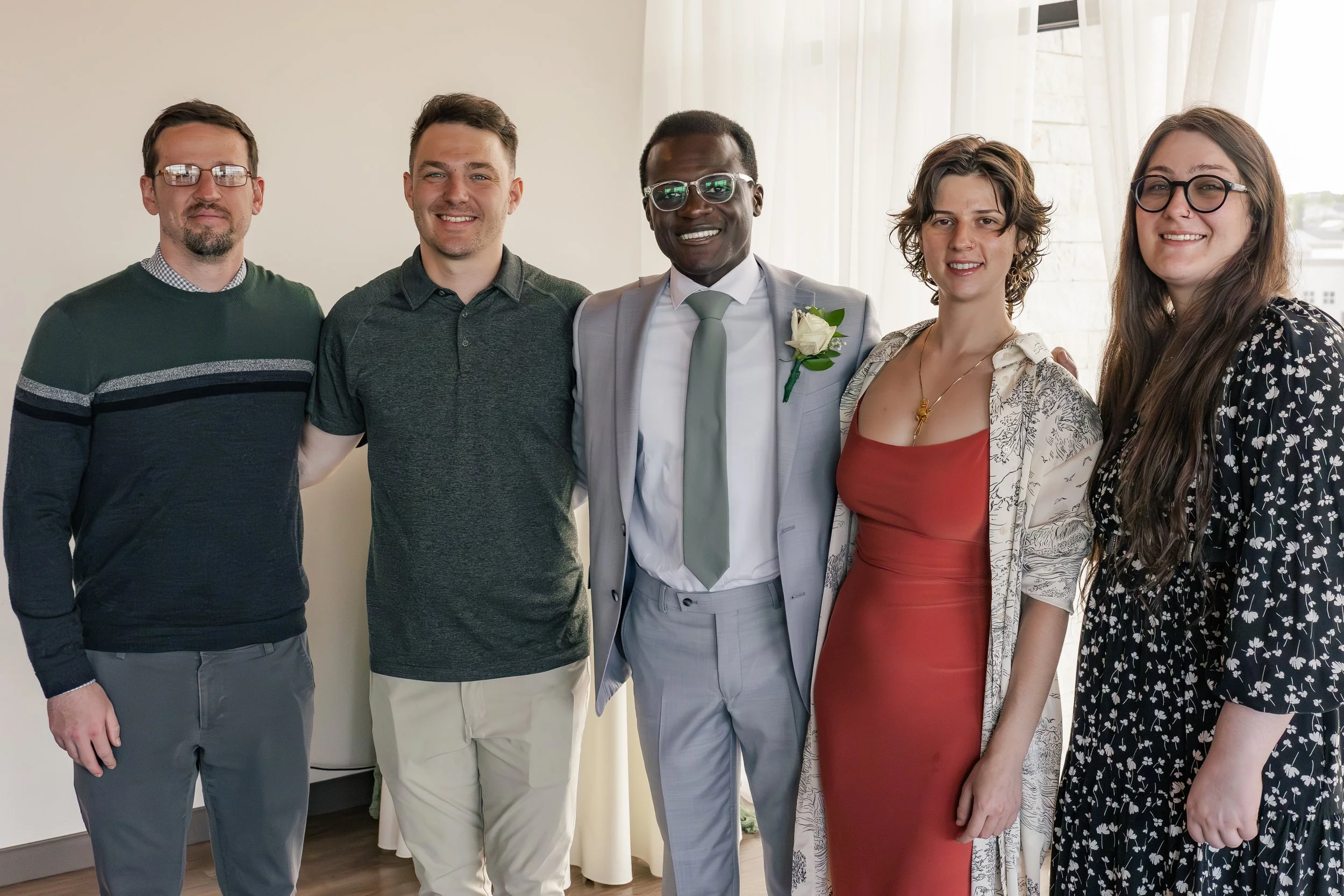 Group of five people, including a man in a gray suit with a white flower boutonniere, posing indoors with a woman in a red dress and floral jacket, and others in casual attire, smiling and standing close together.
