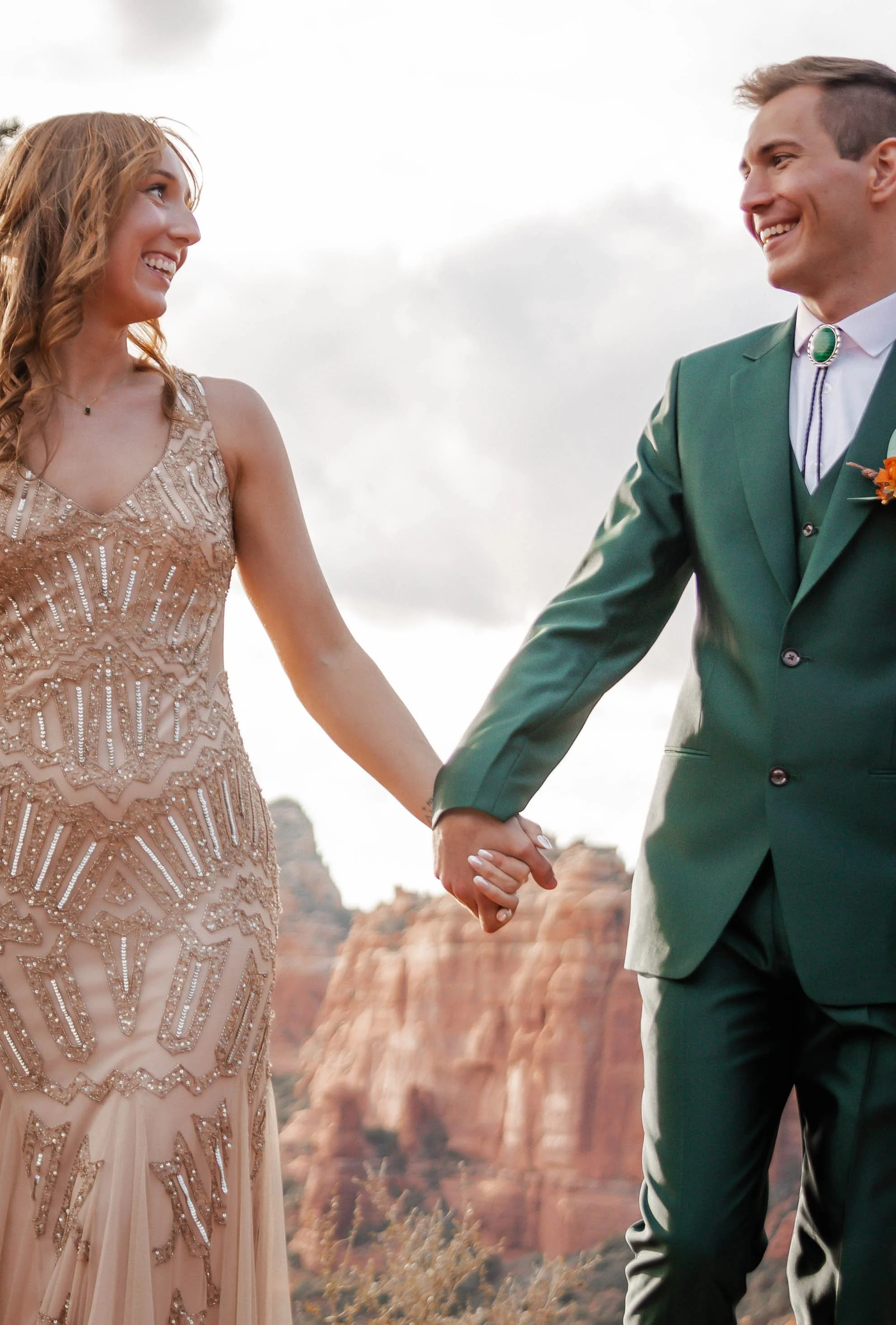 A man and woman holding hands and smiling at each other outdoors, with red rock formations in the background.