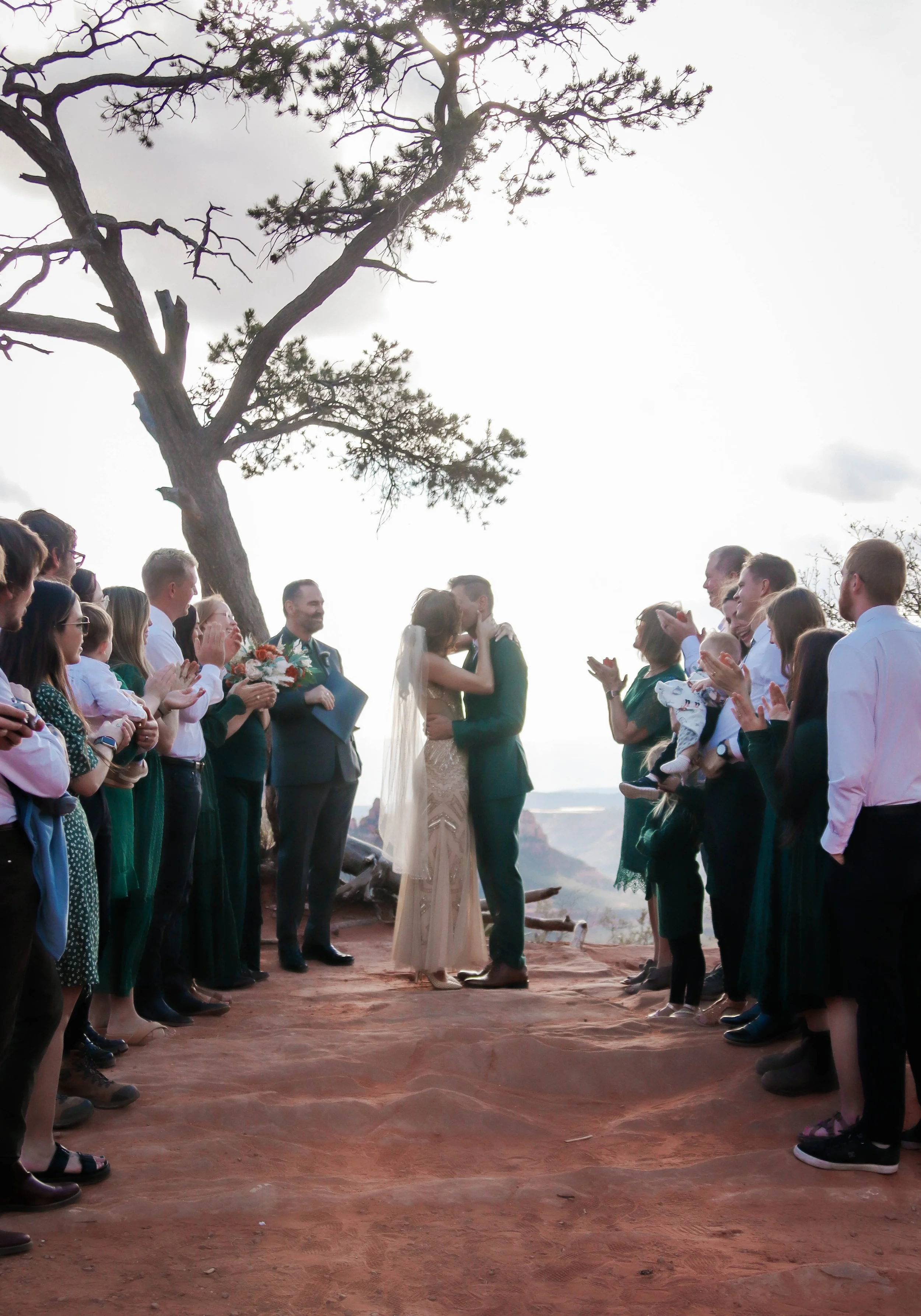 A wedding ceremony outdoors with the bride and groom kissing under a large tree, surrounded by guests clapping and celebrating on a dirt ground with a scenic backdrop.